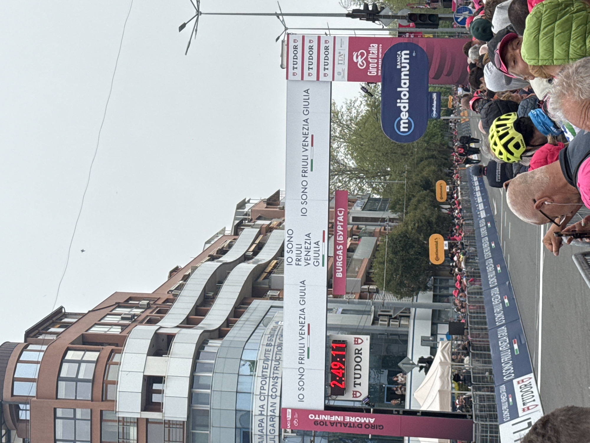 The image shows a scene at the Giro d'Italia cycling race, specifically at a location designated as "Burgas (Бургас)," which indicates a promotional tie-in with the city of Burgas, Bulgaria. A building with modern architecture is visible on the left. The foreground is filled with spectators watching the race, some wearing hats and cycling helmets. Banners promoting the event and sponsors, including "IO SONO FRIULI VENEZIA GIULIA," "TUDOR," "Giro d'Italia," and "mediolanum" are prominently displayed. A digital timer reads "2:49:11," likely indicating the race time. A blue barrier separates the spectators from the race route.