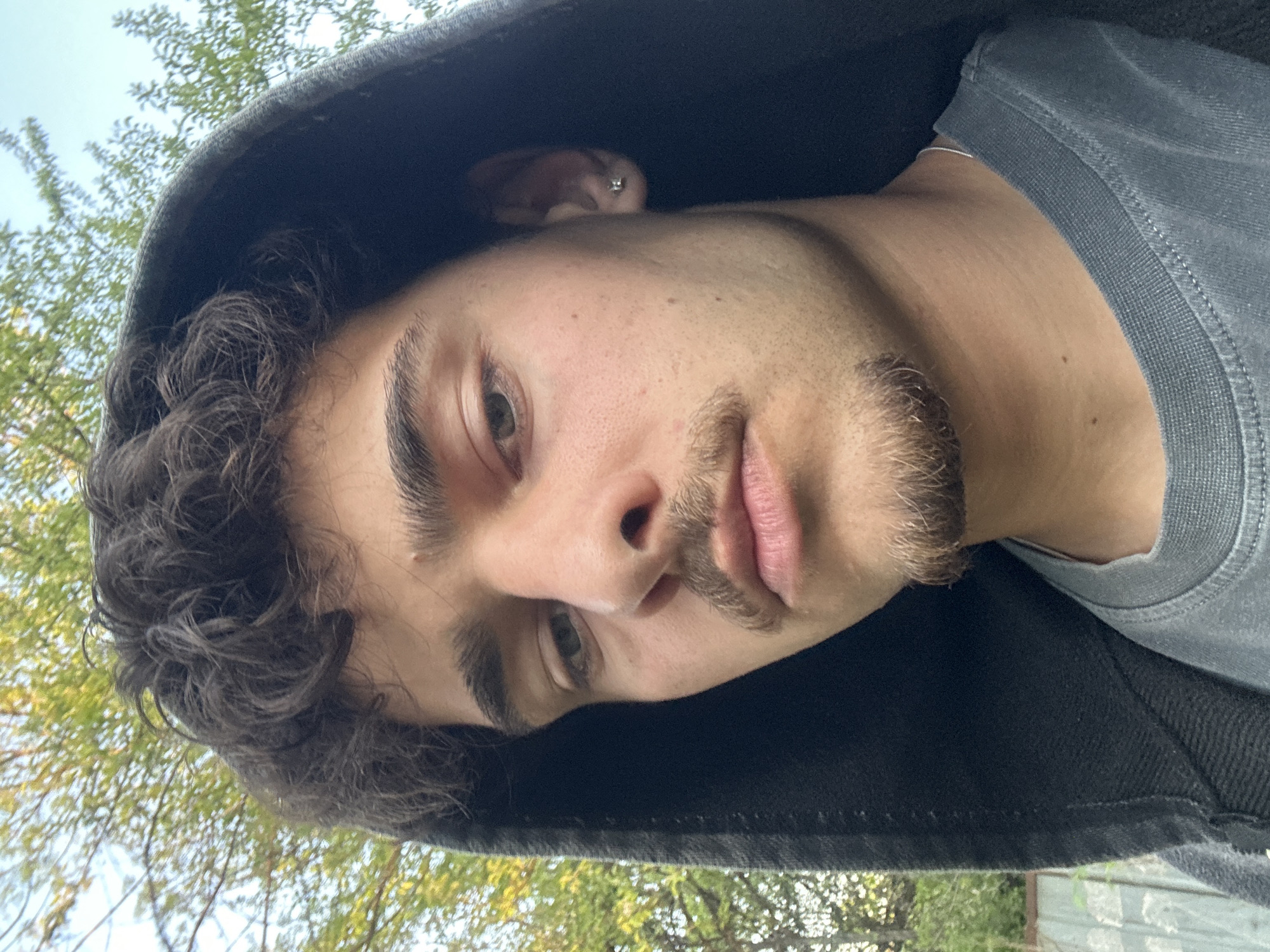 A young man with dark curly hair and a stubbly beard is wearing a dark hooded sweatshirt and a gray t-shirt. He is looking down at the camera with a slight smirk. He has a piercing in his ear. The background is a blurry mix of green trees and a light blue sky. The photo was taken outdoors. There is no definitive information from the photo that places it in Sofia, Bulgaria.
