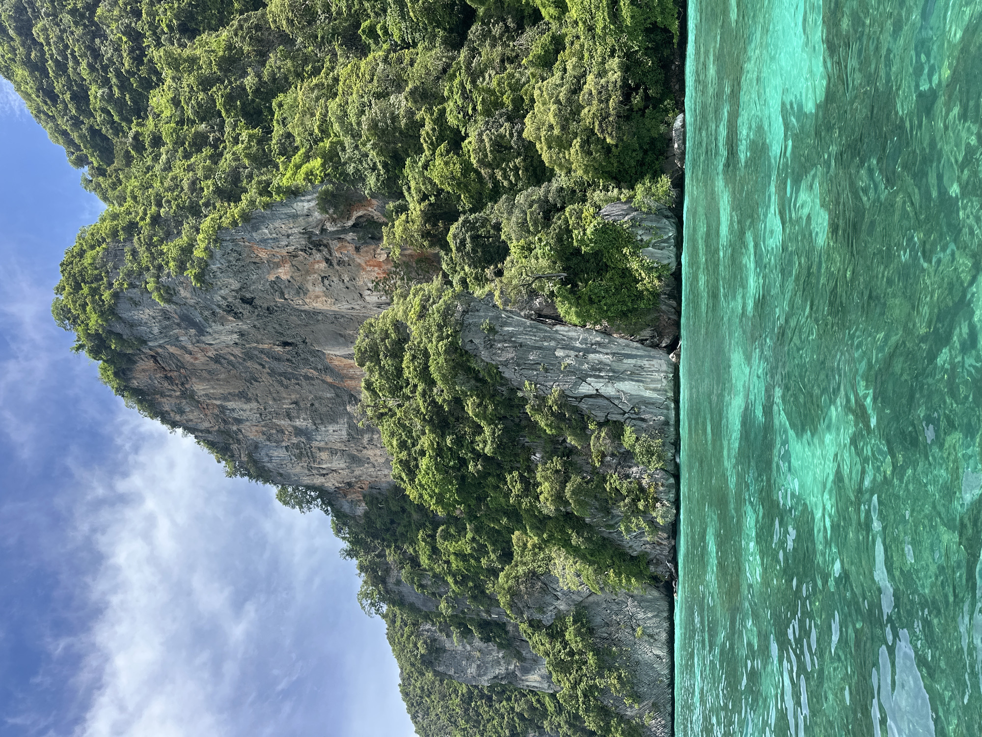 The image shows a dramatic limestone cliff face covered in lush green vegetation, rising from the turquoise waters of the ocean. The cliff is sheer in places, with visible layers of rock, and the vegetation clings precariously to its surface. The water is clear and appears calm, with sunlight glinting off its surface, creating a shimmering effect. The sky above is a bright blue with a few wispy clouds. The perspective suggests the photo was taken from a boat approaching the cliff. The time of day appears to be daytime, likely late morning or early afternoon, given the bright sunlight. The overall impression is one of natural beauty and a sense of adventure. The location is Haad Hin Khom, Thailand, known for its stunning karst landscapes. No people, objects, or text are visible in the image.