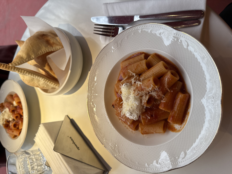 The image captures a top-down view of a dining table set with Italian-style food and tableware, likely in a restaurant setting. The main focus is on a large white plate containing rigatoni pasta covered in a rich red tomato sauce. A generous dollop of white, soft cheese, possibly ricotta or burrata, is placed on top of the pasta, generously sprinkled with grated hard cheese, likely Parmesan, and flecks of black pepper. The plate has an elegant embossed border with a silver or gold rim.
To the left of the main pasta dish, a smaller white bowl holds several golden-brown, triangular pastries or dumplings, possibly empanadas or panzerotti, which are lined with white paper. Further to the left, a smaller plate with a similar pasta dish is partially visible, also topped with white and grated cheese.
In the upper right corner, a knife and fork with dark handles rest neatly on a folded white napkin, awaiting use. Below the main pasta plate, a stack of white paper napkins is held within a dark, possibly metal, triangular holder. The word "operahaus" is visible in white text on the side of this holder. A clear glass with a textured pattern is positioned to the left of the napkin holder.
The table surface is a light, possibly white or cream-colored material, and there's a hint of a red surface or wall in the background on the far left. The lighting in the image appears soft and warm, suggesting an indoor ambiance. The overall presentation is neat and appetizing, indicating a meal in progress or about to be enjoyed.