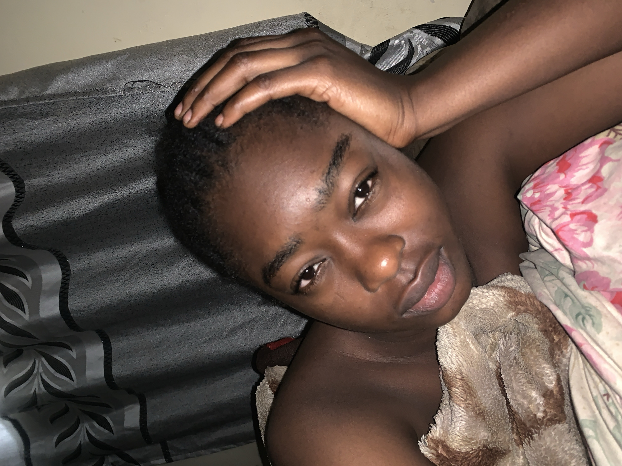 A person with dark skin is lying on a bed with their head resting on their hand. They are looking at the camera. The bed has a gray blanket with a leaf-like pattern and a floral patterned sheet. A brown towel is near their chest. The location is Rafin Par, Nigeria.
