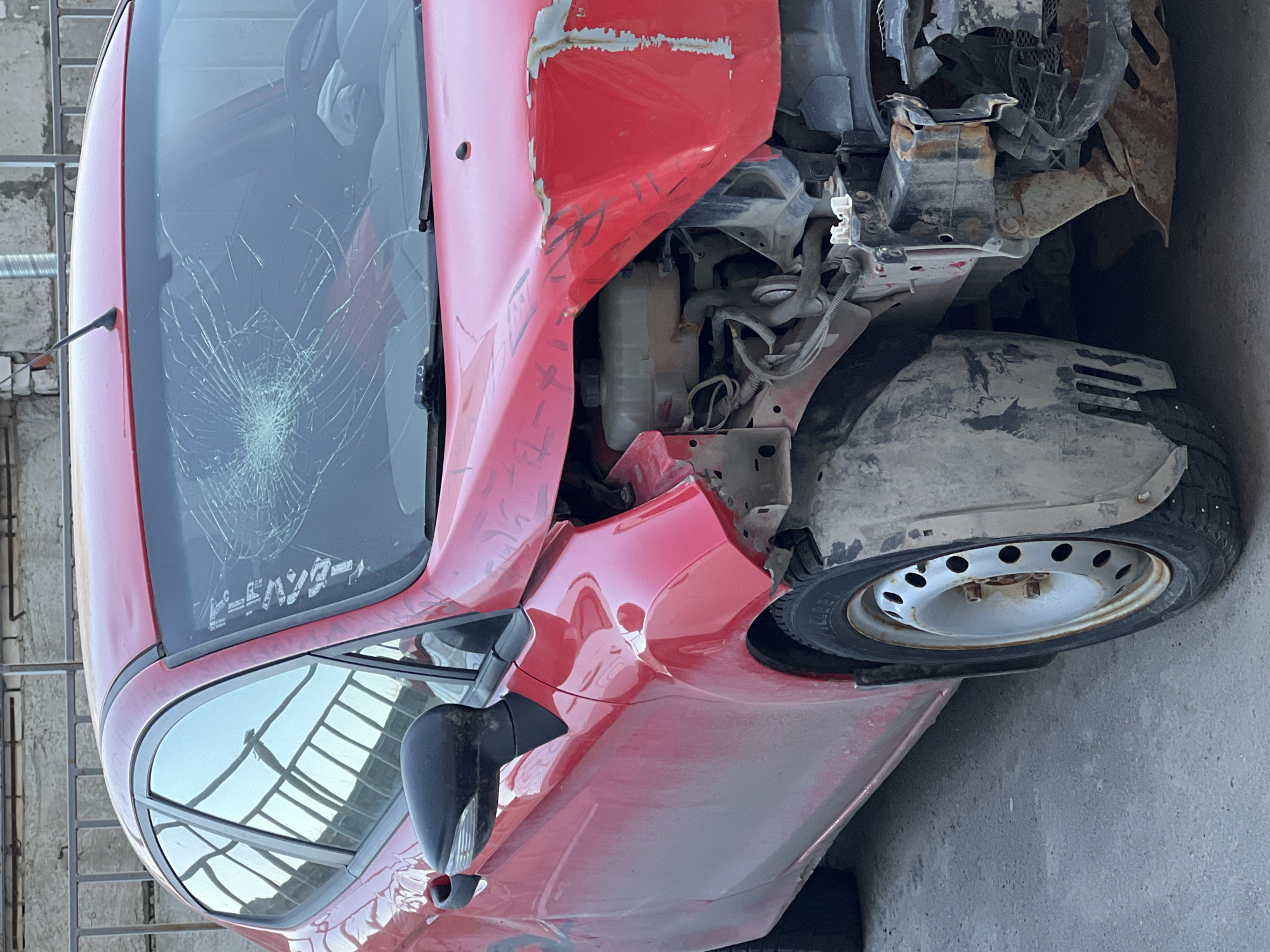A red car is severely damaged, with a shattered windshield and extensive front-end damage, including a missing bumper and exposed internal components. The front passenger-side tire is intact, but the body panels around it are crumpled. The car is likely in Kazan, Russia, as lettering in a Cyrillic-derived alphabet can be seen on the windshield. The environment is a concrete surface, possibly a parking lot or storage area. No people are visible. The scene suggests a collision or other accident.