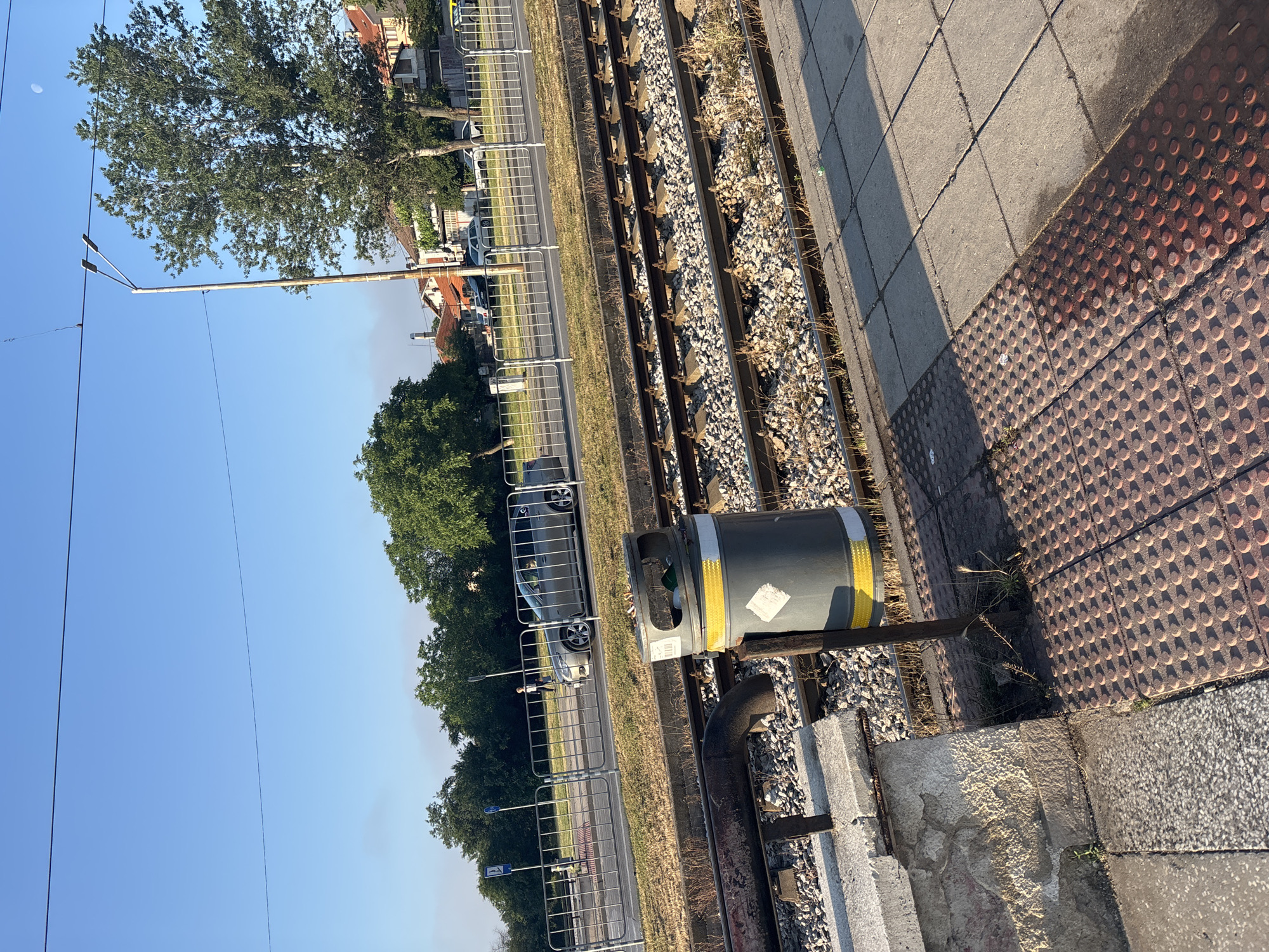 The image depicts a static scene of a railway or tram line in an urban or suburban environment, captured in bright daylight. On the right side of the frame, a concrete platform features both grey square paving stones and a section of red-brown tactile paving with raised dots, indicating a public transport stop. Sunlight casts shadows across the platform, suggesting a low sun angle consistent with morning or late afternoon.
Adjacent to the platform, two sets of railway tracks are visible, with gravel ballast and concrete sleepers. Patches of weeds and dry grass grow between the tracks and along the platform's edge. A grey, cylindrical object, possibly a utility box or a waste bin, stands near the tracks, adorned with yellow reflective tape and an illegible white label. A dark, curved pipe is also present near the tracks in the foreground.
Beyond the tracks, a metal barrier or fence separates the railway area from an adjacent road or pathway. A silver car is clearly visible on this road, with other vehicles and distant buildings with red-tiled roofs forming the background. Green trees with dense foliage line the area behind the fence and buildings.
The upper portion of the image is dominated by a clear, bright blue sky. A faint crescent moon is discernible in the upper left, further hinting at either early morning or late afternoon. A tall utility pole with overhead wires stretches across the sky on the left. A blue sign with a white pictogram resembling a person walking is visible near the fence but lacks legible text. No significant human activity is depicted.
