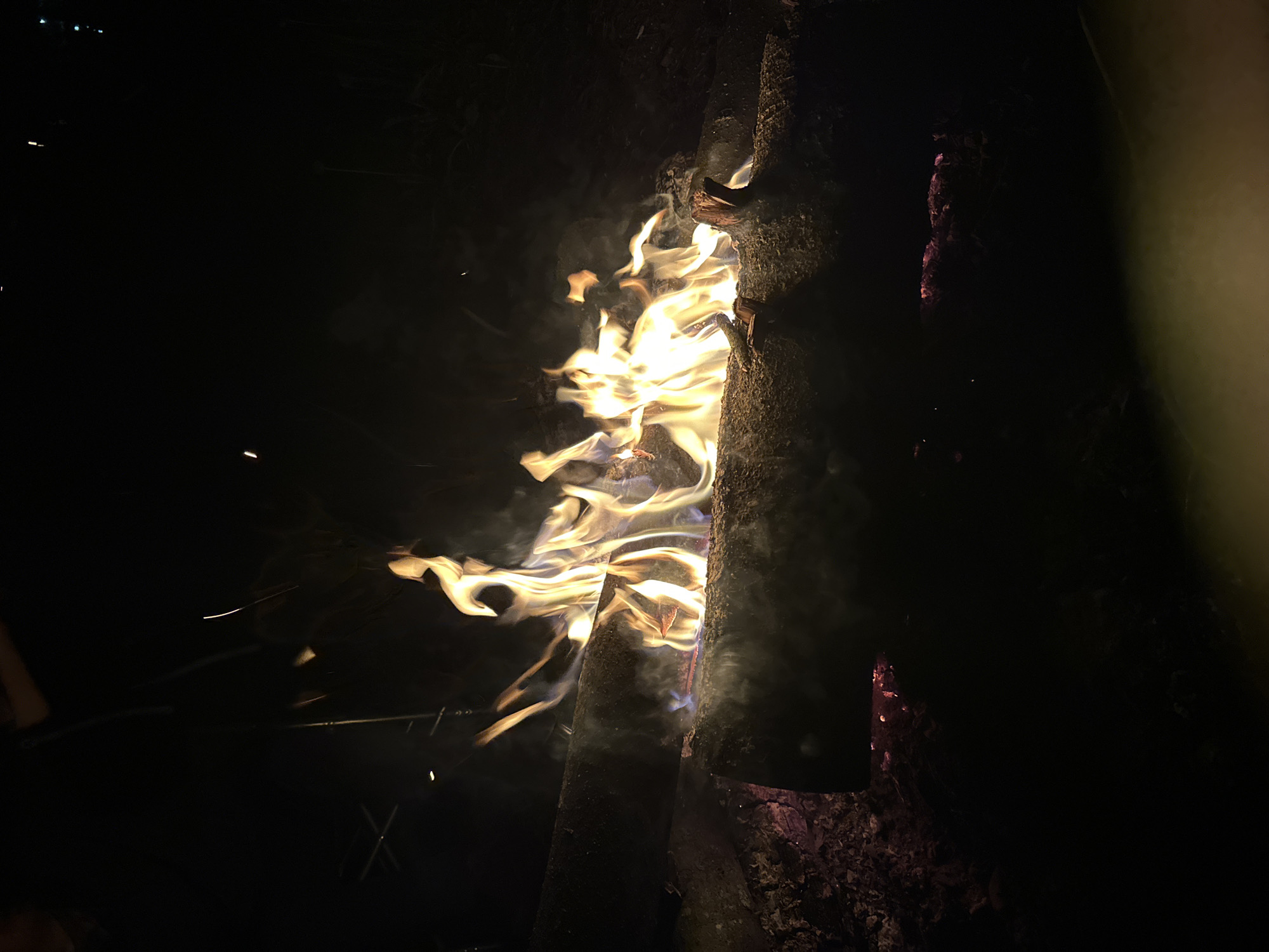 A close-up shot taken at night depicts a campfire consisting of burning logs. The flames are bright yellow and orange, with visible smoke rising. The logs are dark and charred, showing the effects of the fire. Sparks are seen flying off the fire. The surrounding environment is dark. The location, Юзуфоўскі сельскі Савет, Belarus, is not discernible from the image.