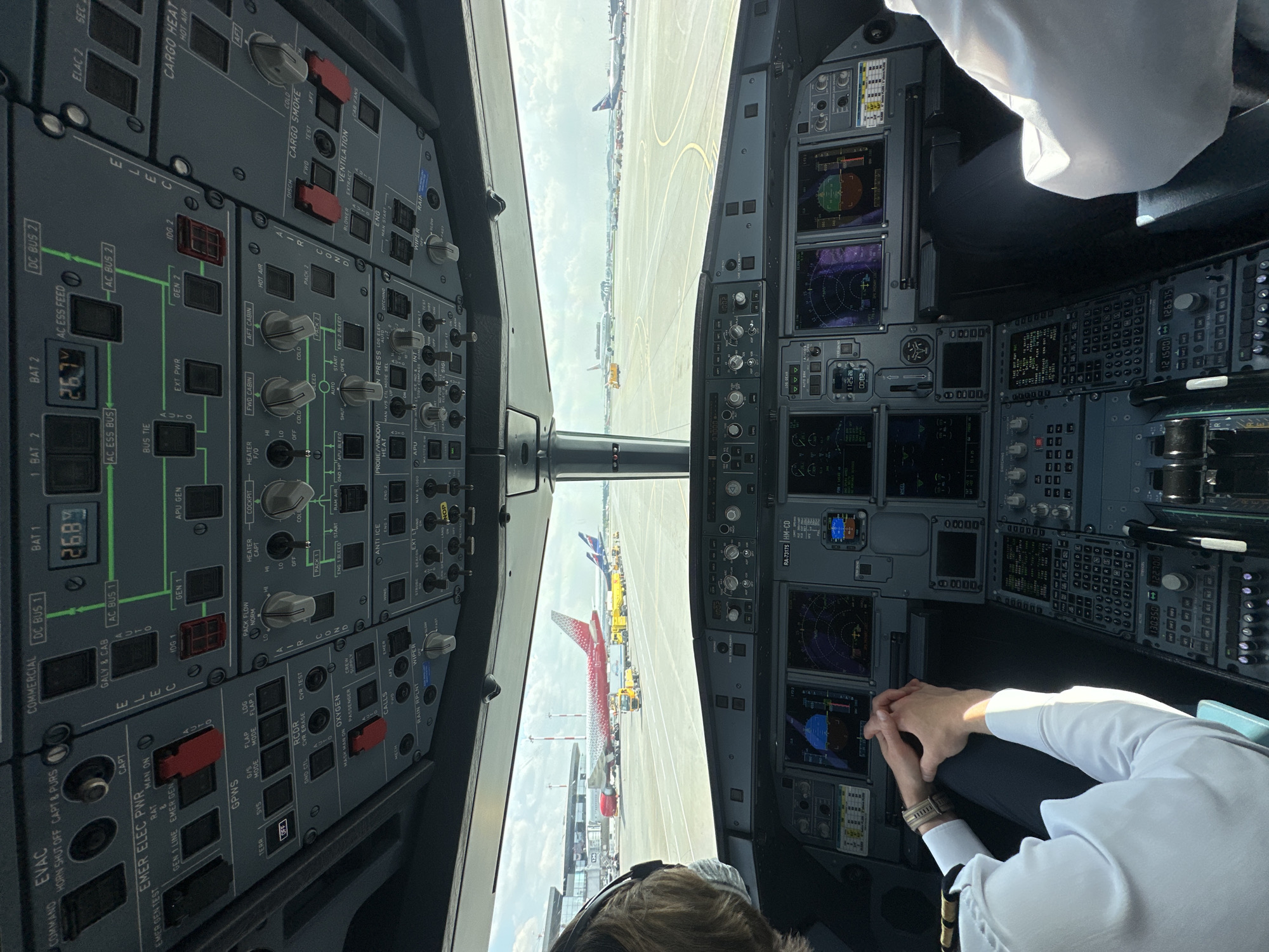 The image depicts the cockpit of an Airbus A320, likely on the ground at Sheremetyevo International Airport, near Khimki, Russia, based on the presence of the Aeroflot Airbus A320, registration number RA-73175, visible outside the cockpit window. Two pilots are visible. The co-pilot is seen from the back in the lower part of the image, while the first officer is visible to the right side of the image. The camera is placed between the two pilots, but slightly closer to the copilot. The primary flight displays, navigation displays, and control panels are visible and illuminated, reflecting the aircraft is powered on. Exterior environmental conditions appear clear and bright. Other aircraft, likely also at Sheremetyevo International Airport, are visible through the windows.
