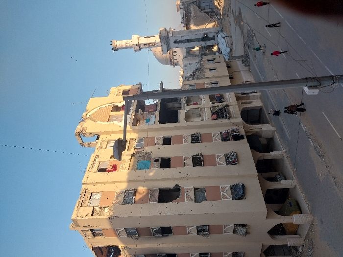 A multi-story building is heavily damaged, with exposed interiors and missing sections of the facade. A mosque with a distinctive minaret is visible in the background, also appearing damaged. Several individuals are walking along a road adjacent to the building. A utility pole stands nearby, partially obscuring the view. The scene conveys a sense of destruction and aftermath.