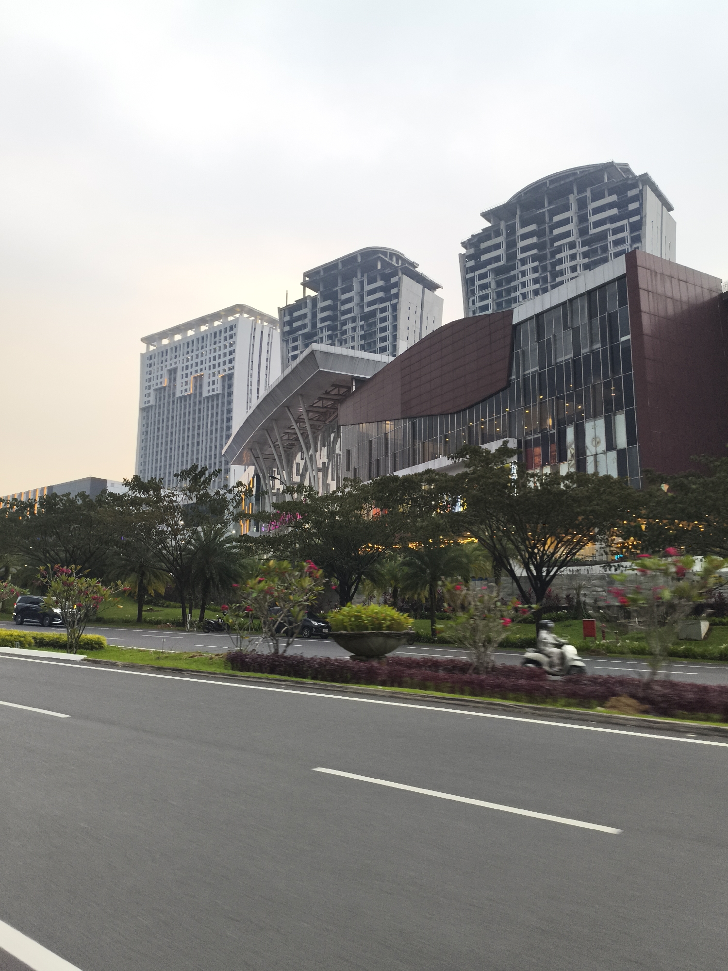 This image depicts a bustling urban scene in Babakan Madang, Indonesia, characterized by modern architecture, a multi-lane road, and lush greenery under an overcast sky. In the foreground, a wide asphalt road stretches forward, marked by distinct white lane lines. A green median strip separates the road, densely planted with mature trees, smaller shrubs, and flowering plants, including visible pink or red blooms and palm trees.
Several vehicles are present, indicating ongoing activity. A black car is partially visible on the left side of the median, while another dark vehicle is further down the road on the right. A white scooter, with a rider wearing a helmet, is captured in motion on the right lane, its blurred form conveying movement.
The background is dominated by a collection of tall, contemporary buildings. A prominent white high-rise with a distinctive grid pattern on its facade stands on the left. Adjacent to it is a large structure, possibly a mall or commercial complex, featuring a dark reddish-brown exterior with extensive glass paneling and a modern, undulating design. Behind these completed structures, several other high-rise buildings appear to be under construction or unfinished, showcasing their gray concrete frameworks against the horizon.
The time of day is difficult to pinpoint precisely due to the hazy or overcast sky, which casts a soft, diffused light over the scene. There's a faint warm, yellowish hue in the sky that could suggest either early morning or late afternoon, or simply a misty atmosphere. No discernible text is visible on any buildings, signs, or vehicles. The scene portrays an active, developing urban environment with a mix of finished and ongoing construction.
