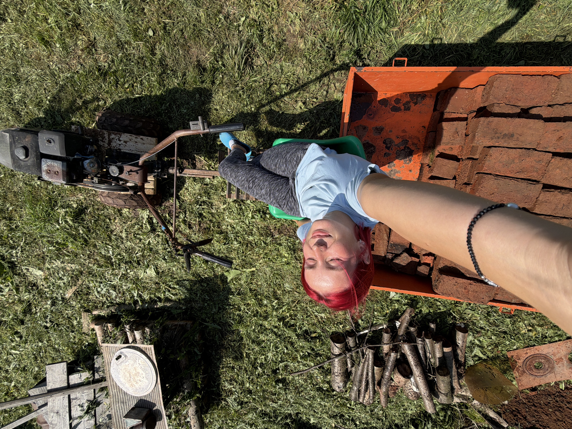 An overhead, high-angle shot captures a young woman with vibrant red hair lying on a green seat attached to an orange, rusted metal cart, which is itself connected to a red and black engine-powered tiller. The woman is reclined with her eyes closed and a relaxed expression, wearing a light blue t-shirt and grey patterned leggings. Her blue clogs are visible. The cart is filled with red bricks. To the left of the cart, a pile of cut logs lies on the grassy ground, next to a wooden structure and a circular metallic object. The scene is set outdoors in a grassy area, with shadows cast by the sun, indicating a daytime setting. The overall environment suggests a rural or suburban yard, possibly in Заболотово, Russia, during warmer weather.