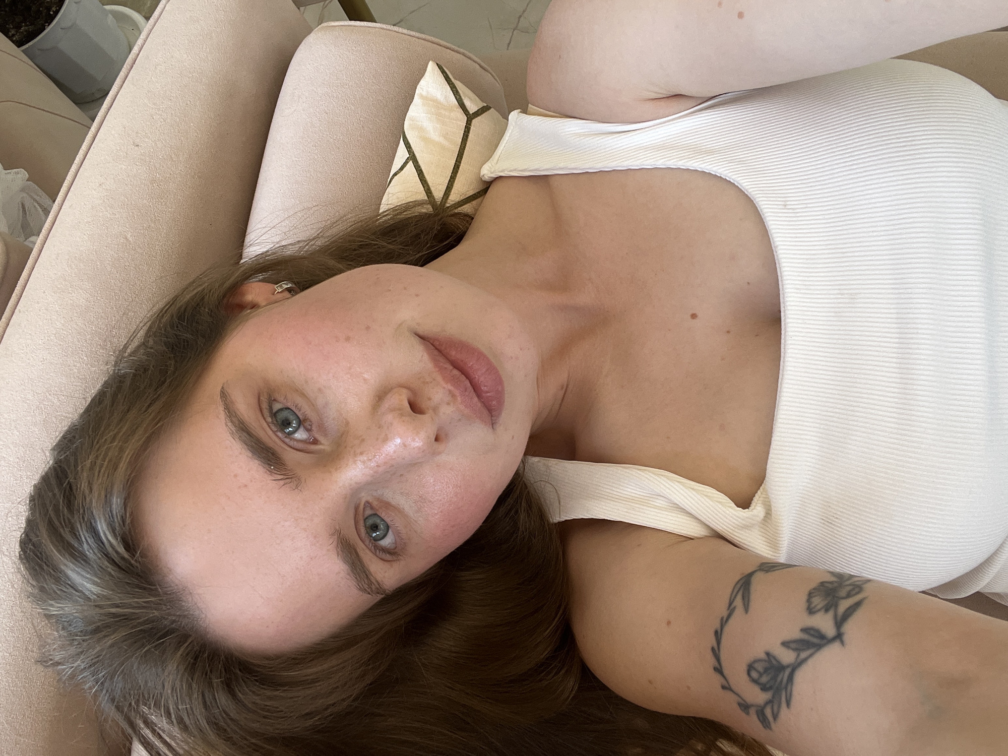 A female subject, appearing to be a young adult, is depicted in an overhead perspective, likely lying on light-colored upholstered furniture indoors. She has medium-brown hair, blue-grey eyes, fair skin with scattered freckles, and visibly flushed cheeks. Her lips are naturally pink and slightly parted. She wears a plain, ribbed, white tank top. A small, silver-toned hoop earring is visible in her right ear. On her left upper arm, a tattoo of a circular floral wreath with leaves and two distinct flowers is present. The background includes a beige or pale pink upholstered surface, consistent with a sofa or armchair, and a small decorative cushion with a geometric pattern in light and dark tones behind her neck. In the upper right corner, a portion of a grey plant pot with a plant is visible. The subject is looking directly into the camera.