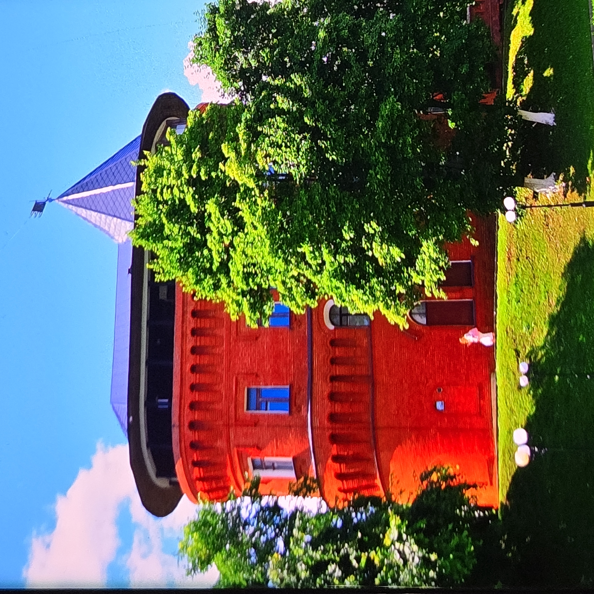 The image shows the Kopylovo Tower in Vladimir, Russia, a cylindrical red brick tower with a conical grey roof topped by a weather vane. The tower is surrounded by lush green trees, and a bright blue sky with scattered white clouds is visible. In the foreground, there is a grassy slope with several lights and a person in a light-colored outfit walking past the tower. The overall scene is that of a historical landmark on a sunny day.