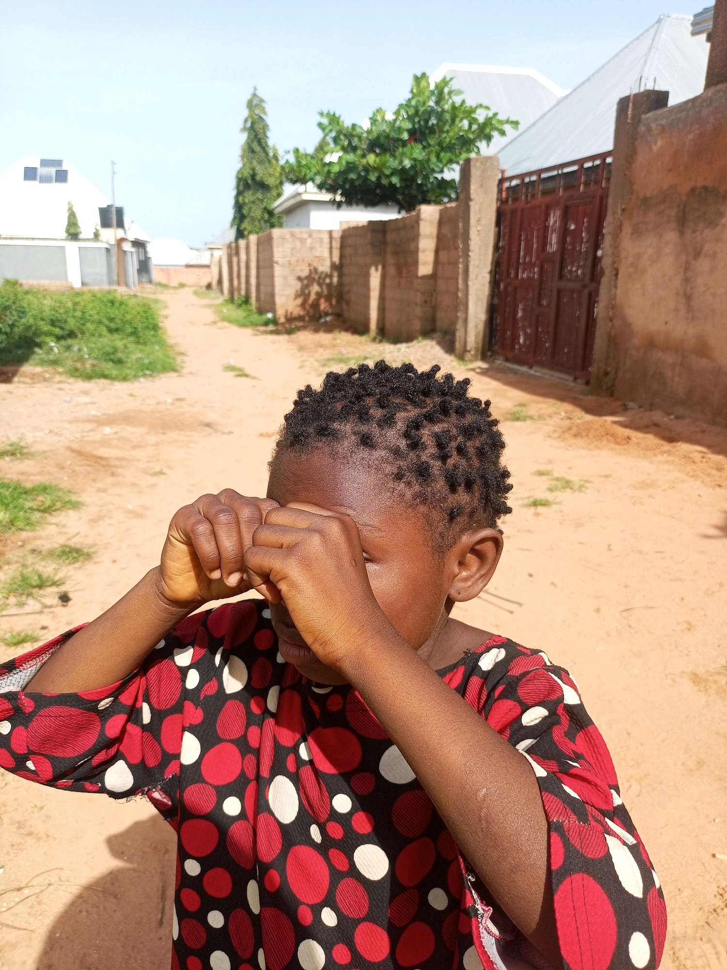 A young Black child with dark, curly hair is standing outdoors in an unpaved street. The child is wearing a red shirt with a pattern of black and white polka dots. Their hands are raised to their face, covering their eyes. The background shows a brick wall, a metal gate, a tree, and buildings. The ground is sandy and there is some dry grass on the sides of the street. The sky is clear and blue. The location appears to be a residential area in a town in Nigeria.