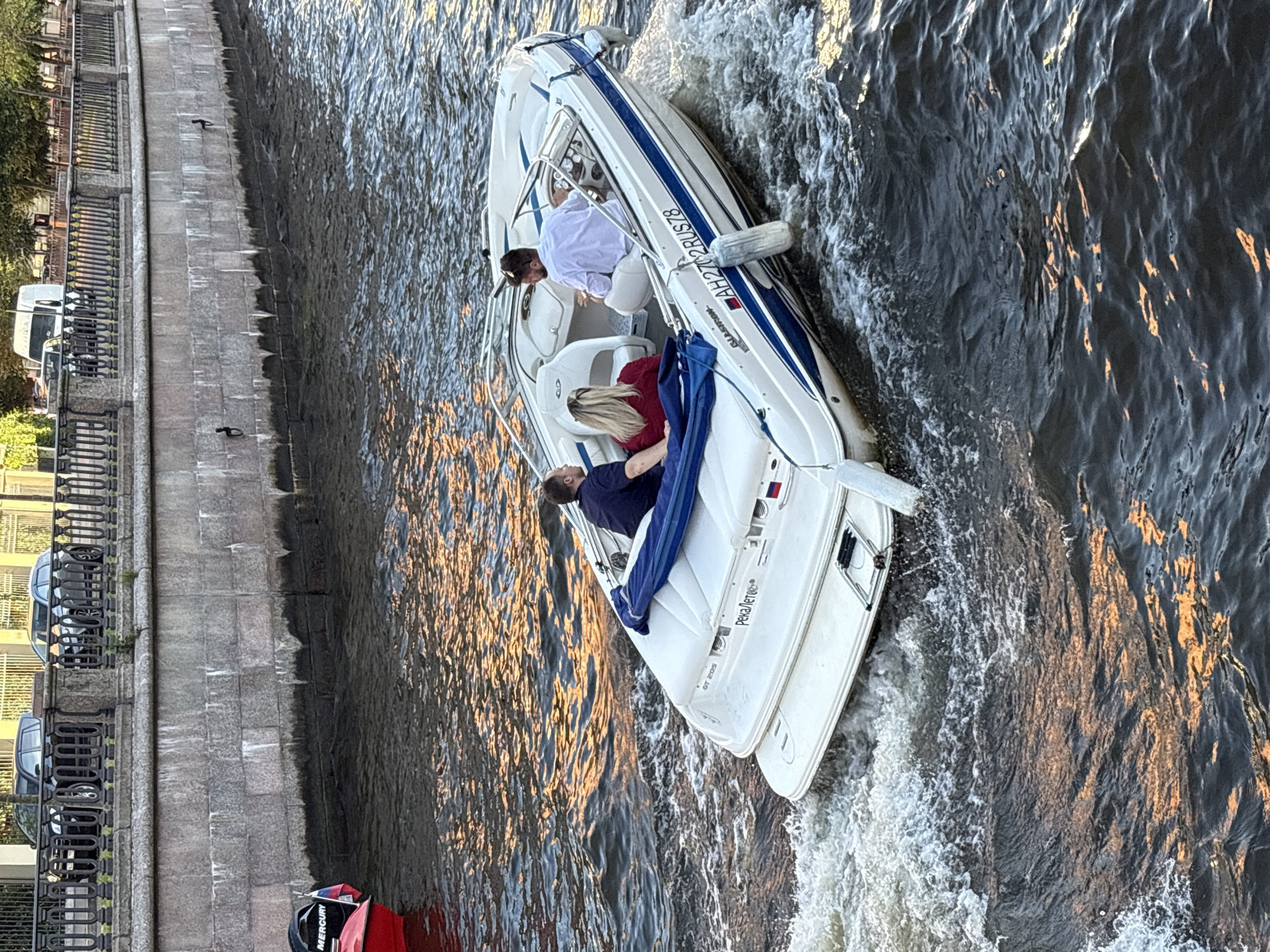 A white motorboat with blue trim is moving through dark water, leaving a foamy wake. Three people are on board: two men and one woman. One man, wearing a white shirt, is steering the boat from the right seat. Another man, in a dark blue shirt, sits in the back left. A woman with blonde hair, wearing a red shirt, is seated between them. The boat has Russian registration numbers AH2702RUS78. In the background, a stone embankment lines the canal. On top of the embankment, a decorative metal fence is visible, with parked cars and a white van behind it. Trees and buildings are further in the distance. A red boat with "MERCURY" branding and a Russian flag is partially visible on the left edge of the frame. The water reflects the warm glow of what appears to be a setting sun or artificial lighting. The scene is likely taking place on one of the many canals or rivers within Saint Petersburg, Russia.