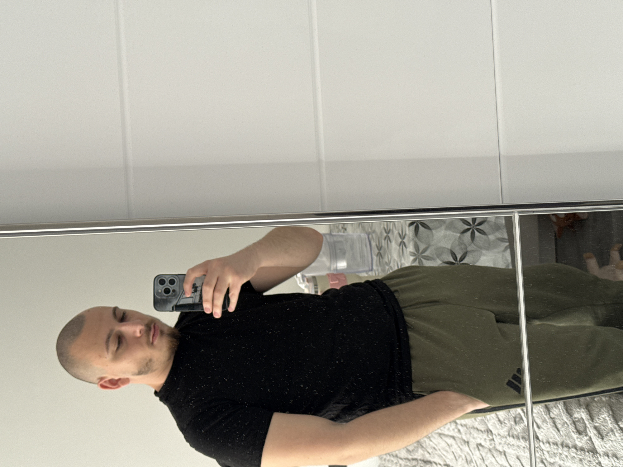 A bald man wearing a black t-shirt and green Adidas sweatpants is taking a selfie in a mirror. The background of the mirror shows a white tiled wall, a water bottle, patterned wallpaper, and a blanket. The location is likely indoors, but the city of Stara Zagora, Bulgaria, cannot be determined from the image.