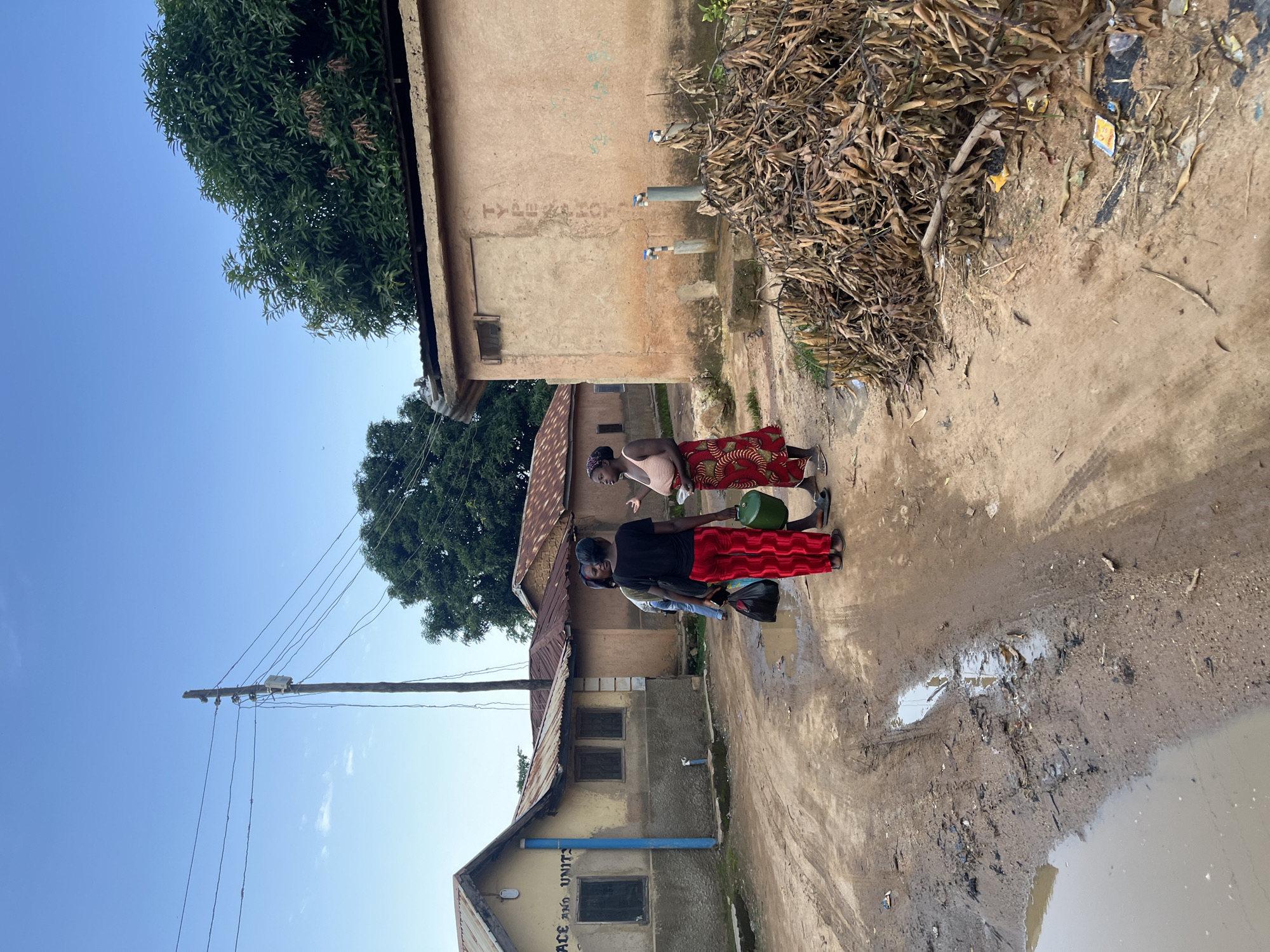 The image depicts an outdoor scene in Rafin Par, Nigeria, featuring three individuals standing on a muddy ground next to a building. Two of the individuals are women, one wearing a red and white patterned skirt and the other a red patterned skirt and a black shirt. The third person appears to be a child. The woman in the red and white skirt is holding a green container, suggesting they are collecting water.
The building behind them is made of light-colored plaster and has a corrugated metal roof. Two faucets are visible on the wall of the building, indicating a water source. There are also piles of dried leaves and debris near the building. The ground is uneven and wet, with puddles of water, hinting at recent rainfall or a consistent water issue. A wooden utility pole with electrical wires stretches across the sky, suggesting the presence of electricity in the area.
The sky is clear and blue, indicating a sunny day, likely in the late afternoon or early morning due to the angle of the light. The overall atmosphere appears to be one of daily life activities, potentially involving the collection of essential resources. There is some faint text on the building, though it is difficult to discern clearly. One visible segment on a separate building reads "PEACE AND UNITY". The scene appears to be a rural or semi-rural setting.