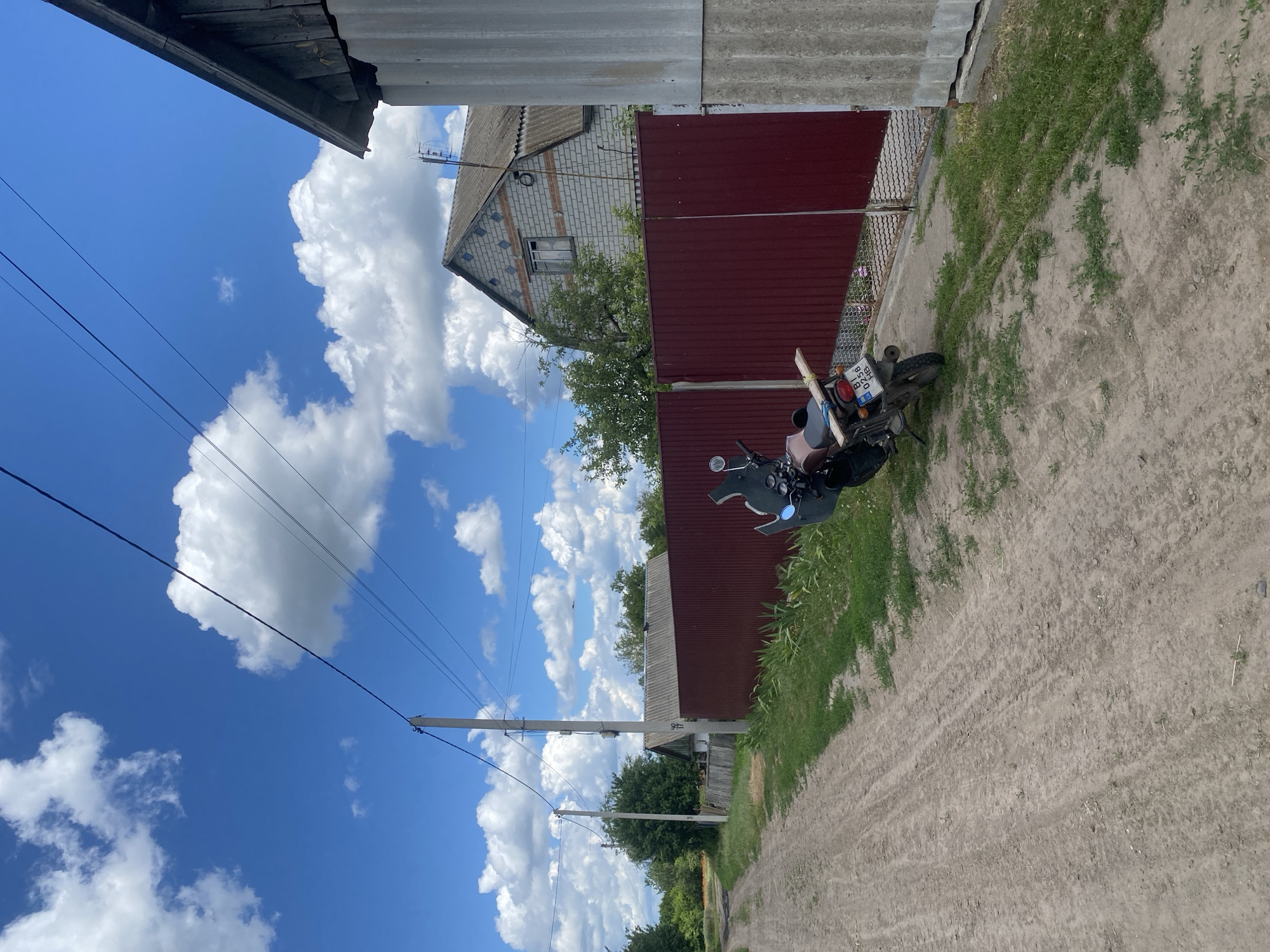 This image depicts a rural scene on a sunny day with scattered white clouds against a blue sky. In the foreground, a dirt road with tire tracks leads into the distance. To the right of the road, a red metal fence encloses a property. A parked motorcycle with a license plate reading "BI 0258 HB" is situated by the fence. A light brown wooden board is resting on the rear of the motorcycle. Beyond the fence, a house constructed of white brick with orange accents and a grey roof is visible. A tall utility pole with power lines stretches across the upper portion of the frame. The environment suggests a residential area in the village of Ustyvytsia, Ukraine, characterized by simple structures and unpaved roads.