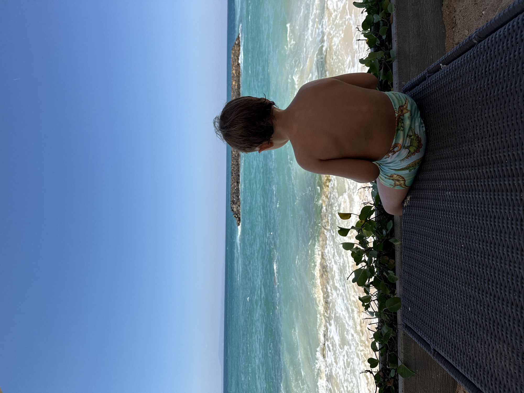 A young child, observed from a rear perspective, sits on a dark grey, woven lounge chair situated on sandy ground. The child has short, dark brown hair and is shirtless, wearing light blue swim shorts adorned with green dinosaur patterns. The child faces away from the camera, gazing towards the ocean. A low concrete barrier topped with green, leafy plants separates the lounge chair from the beach. Beyond the barrier, white-capped waves break on a light sand beach, leading into clear turquoise-to-blue ocean waters. A dark rock barrier or breakwater is visible in the mid-distance ocean to the right. The horizon line is clear, with a faint, distant landmass visible to the left. The sky is a cloudless, vibrant blue, with a single, faint white contrail visible high overhead. This scene unfolds on a sunny day in Kuta Selatan, Indonesia.