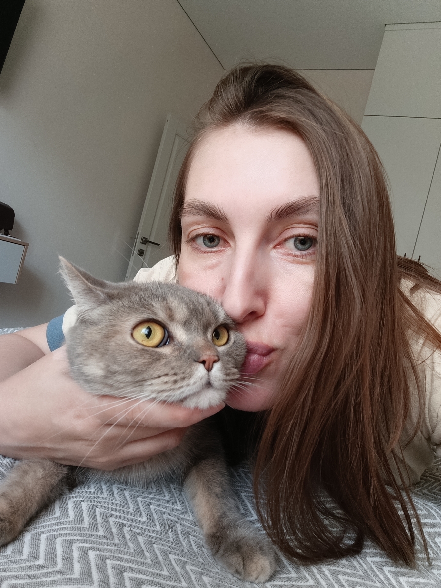 A woman with long, brown hair lies on a gray and white patterned surface, possibly a bed. She is holding a gray tabby cat with large, yellow eyes close to her face, appearing to give it a kiss. The location is Chaikovsky, Russia. The background includes a white door and light gray walls. There is a small, white shelf with a dark object on it visible in the upper left.