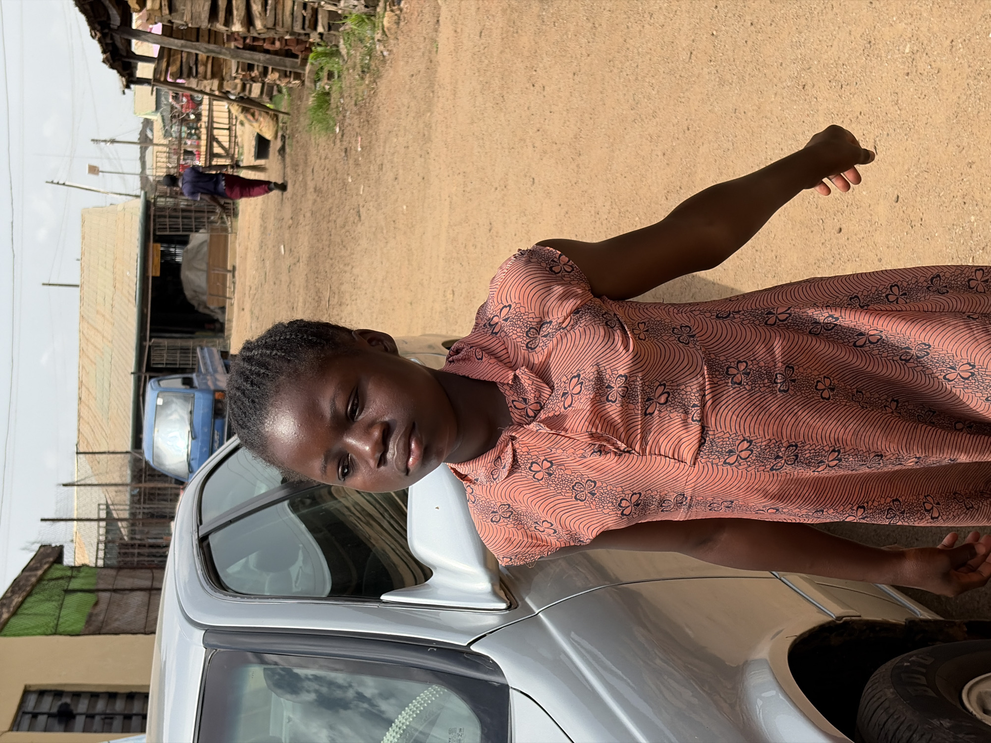 A young woman with braided hair wearing a peach-colored floral print dress stands next to a silver car. Her right arm is raised. She is looking towards the camera. The location is Jos, Nigeria, suggested by the buildings in the background. A pedestrian wearing dark clothing is walking away from the camera in the background, in what appears to be a sandy, unpaved street. Wooden structures and corrugated metal sheeting are visible, indicating a residential or commercial area. A blue vehicle is parked nearby. Tires can be seen near the silver car.