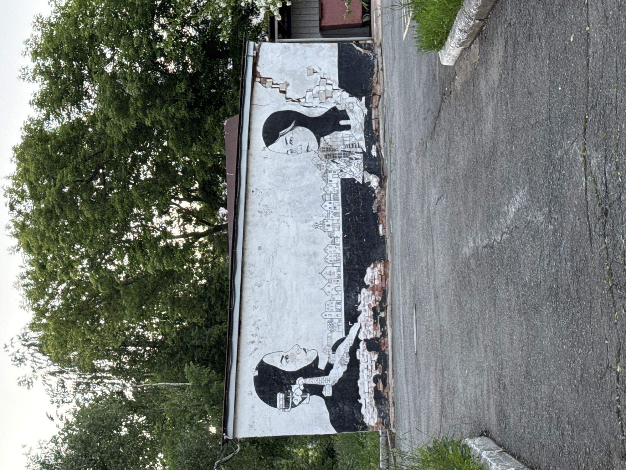 The image depicts a mural painted on the exterior wall of a building. The mural features two portraits of women in profile, facing each other. The woman on the left has a black headscarf and headphones, with imagery of buildings and industrial elements emanating from her head. The woman on the right has a similar headscarf and a more minimalist depiction of a cityscape behind her. The mural is rendered in black and white and appears to be on a weathered wall with visible brickwork where the plaster has crumbled. Behind the building, lush green trees are visible against a bright sky. In the foreground, a paved asphalt surface extends towards the building, with a concrete curb and grass on the sides. The overall impression is one of urban art situated in an outdoor, possibly industrial or semi-abandoned, setting.