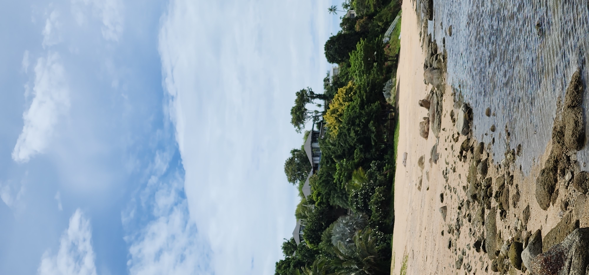 A tropical beach scene unfolds under a partly cloudy sky in Ko Samui District, Thailand. The foreground is dominated by a sandy beach littered with numerous grey and brown rocks of various sizes. Some of these rocks are partially submerged in the clear, rippling water that extends to the right of the frame. The water exhibits a blue-grey hue.
Further inland, the beach transitions to a lush green hillside covered in dense vegetation. Several modern villas with dark grey roofs are visible nestled amongst the trees on the upper slope. Palm trees and other tropical flora contribute to the vibrant greenery. The sky above is a light blue, with scattered white clouds. The overall impression is one of a serene coastal landscape.