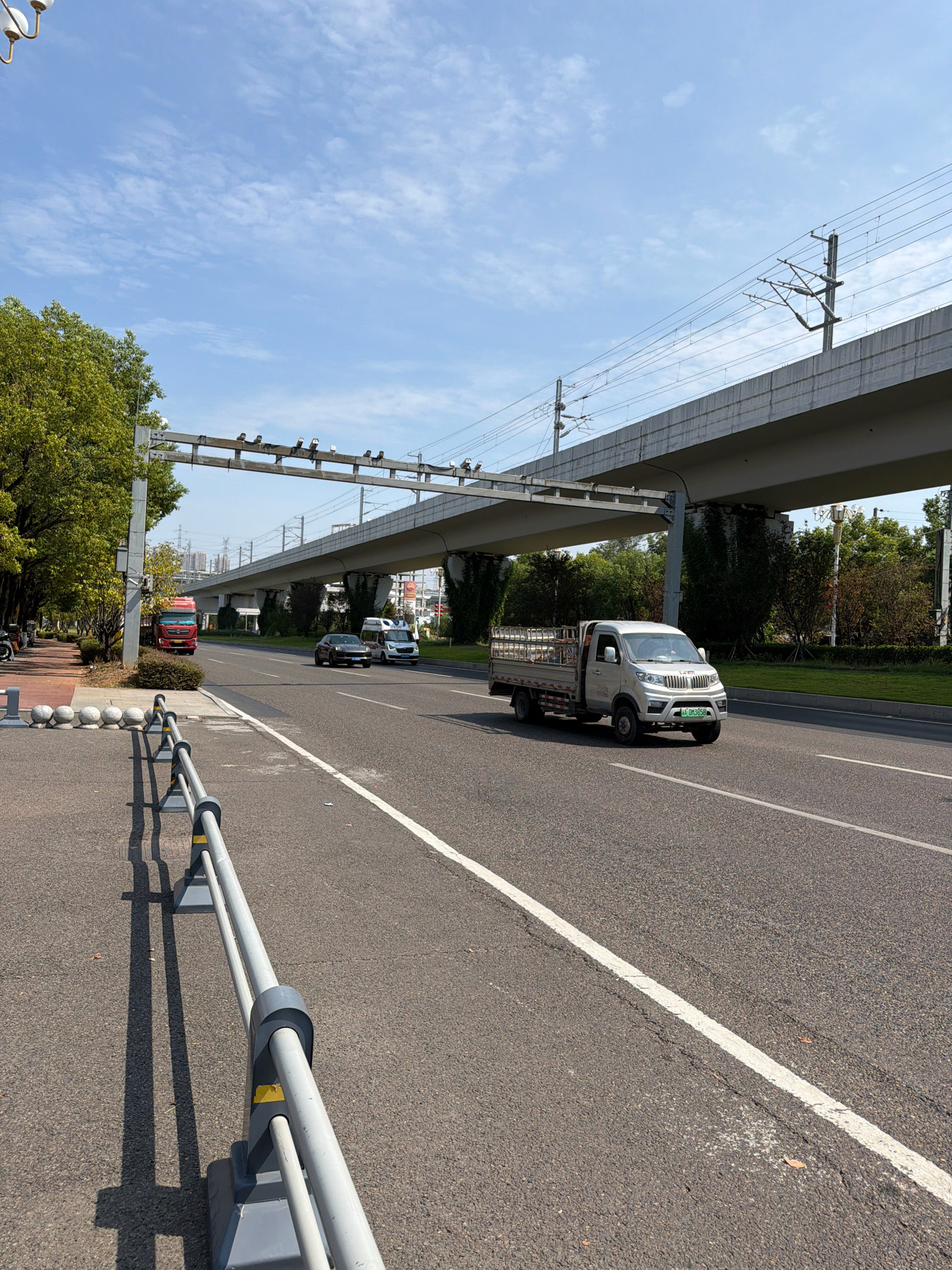 The image shows a sunny day with a blue sky and scattered clouds. In the foreground, a grey asphalt road with a white line marking runs diagonally from the bottom right to the upper left. A grey metal barrier with yellow accents is positioned along the left side of the road.
In the middle ground, several vehicles are on the road. A white truck with a metal cage in the back is driving in the right lane. To its left, a dark sedan is visible, and further left, a white van. In the left lane, a red truck is parked. A sidewalk with spherical bollards is to the left of the parked truck, lined with trees.
Overhead, a modern elevated structure, likely part of a transportation network, spans across the scene. This structure appears to be a viaduct or bridge for a railway or elevated highway, with visible overhead power lines and supporting pylons. Greenery, including trees and ivy, covers parts of the supporting structures and the surrounding landscape. In the distance, city buildings are visible. The overall impression is an urban environment with active transportation infrastructure. This scene is located in the city of Jiangjin, China.