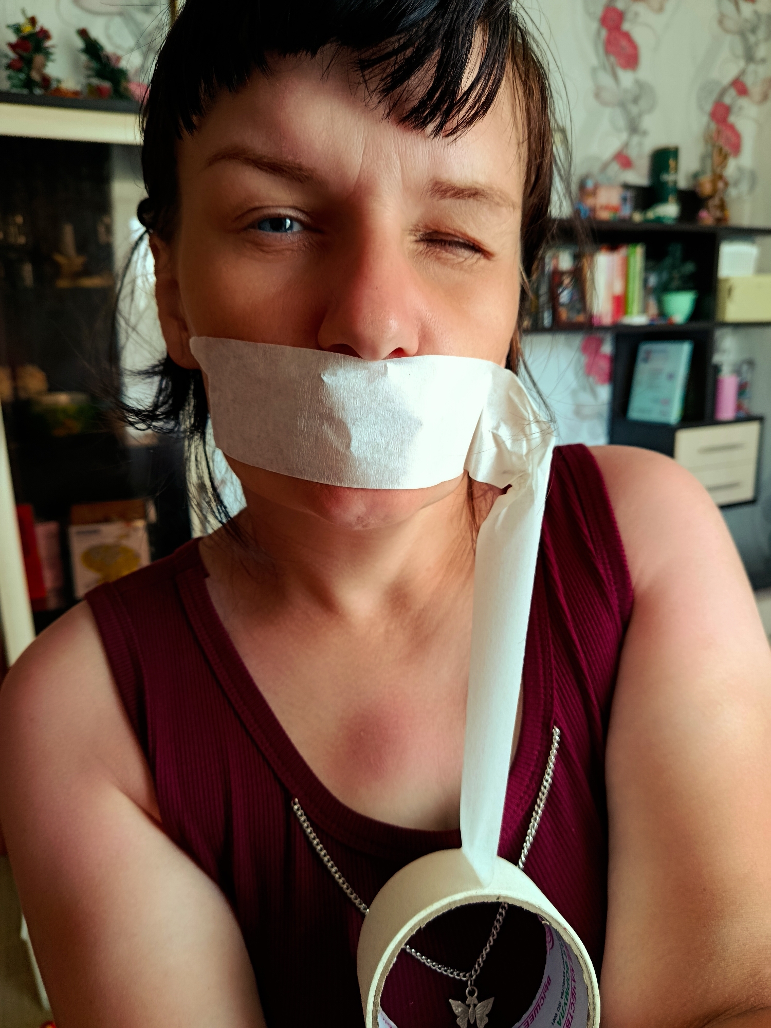 A medium, eye-level shot depicts a woman with short, dark hair and bangs, wearing a maroon tank top with a delicate silver chain necklace featuring a butterfly charm. A strip of white paper, possibly from a roll of tape visible in the lower right corner of the frame, is taped across her mouth. One eye is closed in a wink, while the other eye is blue and open. The background shows a bookcase with various items, including books, plants, and decorative objects. The wallpaper behind the bookcase has a floral pattern. The location is Фоки, Russia.
