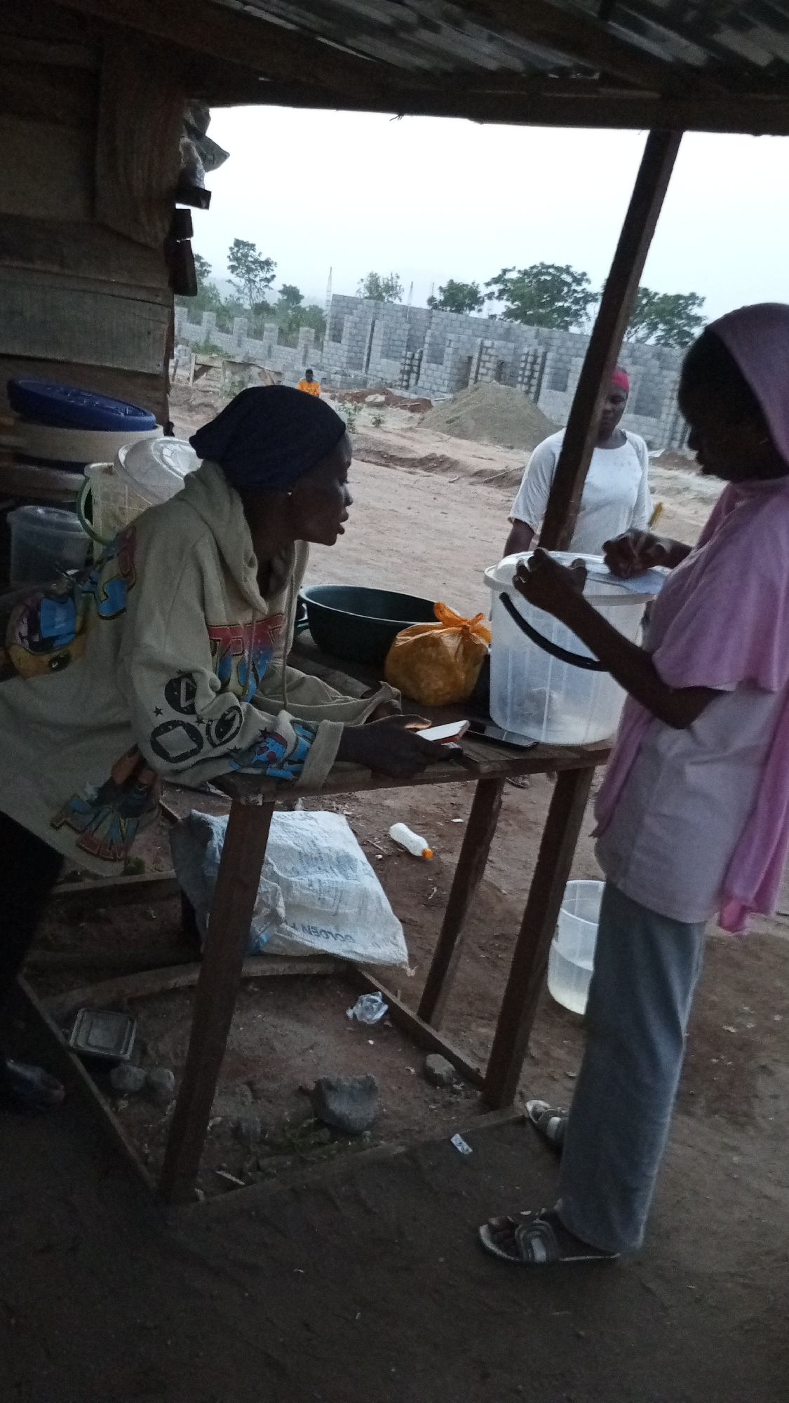 In Adankolo, Nigeria, the image captures a scene under the shelter of a small, rudimentary wooden structure. Two women stand on a dirt ground surface, adjacent to a wooden table cluttered with various items: containers, a cooking pot, and an orange plastic bag. One woman, facing left, is dark-skinned and is wearing a hooded sweatshirt with colorful designs and a dark headscarf. She appears to be holding some papers. The second woman, seen partially from the side, is wearing a pink headscarf and a long-sleeved shirt, and is handling a clear plastic bucket. A third woman in a white shirt is visible in the background. Behind them, a construction site is apparent, featuring walls in the process of being built and piles of sand. Other items visible include a sack with the word "GOLDEN" on it, a plastic bottle, and more plastic containers stacked near the roof of the shelter. The lighting suggests it is either early morning or late afternoon.