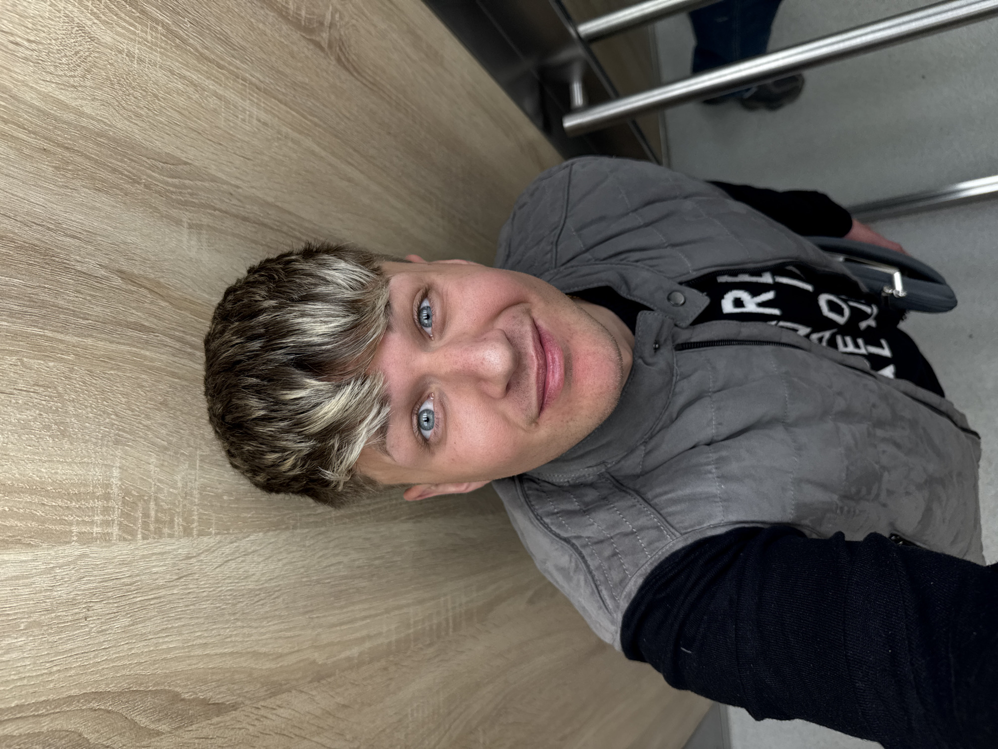 A medium shot depicts a man with fair skin, short gray hair with blonde highlights, and blue eyes. He is smiling slightly. He is wearing a gray quilted vest over a black top with white text. He appears to be indoors, with a light wood-toned surface on the left, and metal railings with a person in blue jeans in the background. The location is not identifiable as Plovdiv, Bulgaria, from the given visual information.