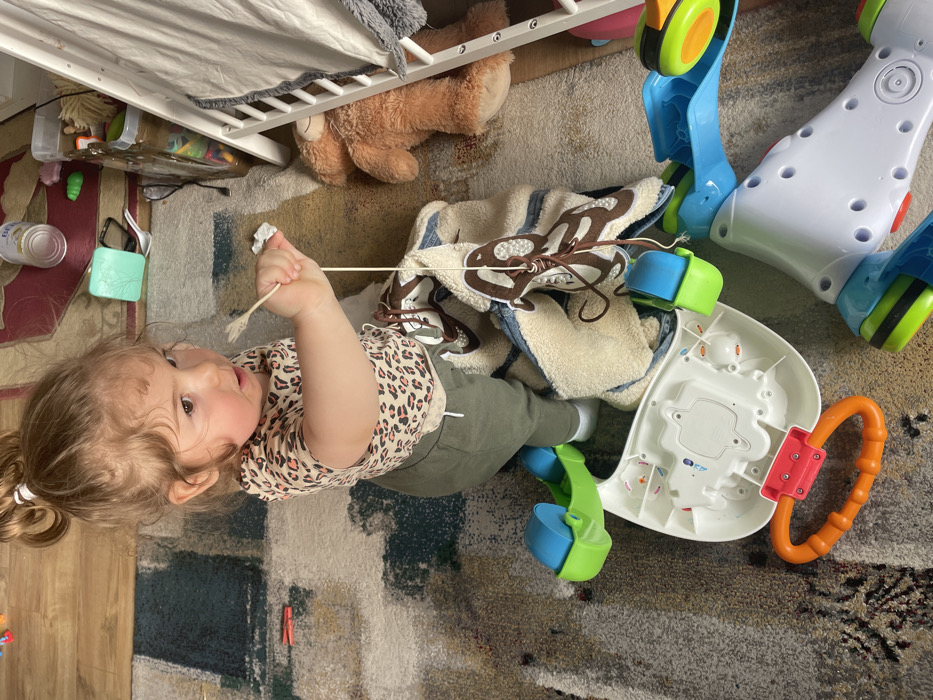The image displays an indoor scene featuring a young child on a patterned carpet. The specific location in Senden, Germany, cannot be confirmed from the visual information provided.
A toddler, with light brown, curly hair secured in a high bun with a white elastic band, is visible in the center. The child wears a light-colored t-shirt with a brown leopard print pattern and olive-green pants, with white socks visible on the feet. The child is oriented slightly upwards and to the right, exhibiting an open-mouthed expression. The child's right hand grips a thin, light-colored stick-like object, possibly a skewer, alongside a small piece of white material. A thin string or cord descends from the stick, attaching to an off-white, textured blanket with brown, monkey-like patterns, which is partially draped over a toy.
The foreground and midground on the carpet contain several objects: an inverted white plastic toy base with attached blue and green components, another blue and green plastic toy piece, and an orange ring-shaped teether or handle with a red connector. To the right of the child, a brown teddy bear lies on its back adjacent to a white wooden structure, possibly a bed frame or doll's bed, partially covered by white fabric.
The background reveals a section of light brown wooden flooring and darker carpet, upon which various items are scattered, including a silver can, a light green plastic container, a white plastic fork, a red plastic clothespin, black-framed glasses, and a black cable. The floor throughout the visible area is covered by a multi-colored carpet featuring abstract patterns in shades of beige, grey, blue, and green. The scene is illuminated by ambient light.