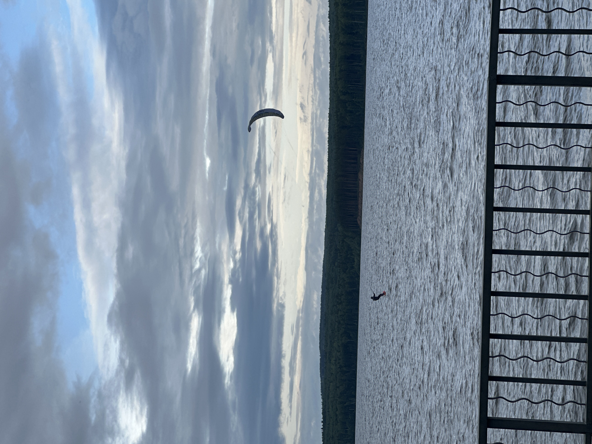The image depicts an outdoor scene located in the city of Chaikovsky, Russia, featuring a vast body of water under a partially overcast sky. The upper two-thirds of the frame are dominated by a sky filled with dense grey cloud cover, exhibiting varying light and shadow, with intermittent patches of lighter blue sky visible. Below the sky, a distant horizon line reveals a continuous stretch of dark green forest, suggesting a forested landmass across the water.
On the surface of the dark blue-grey water, which shows discernible ripples indicating wind activity, a lone individual is engaged in kitefoiling. The person, dressed in dark clothing, is standing on a board equipped with a hydrofoil mast, which elevates the board and rider above the water. High in the mid-right section of the sky, a large, crescent-shaped kite, predominantly white and dark in color, is visible, connected by faint lines to the individual on the water, providing propulsion.
The immediate foreground of the image is occupied by a dark metal railing, composed of vertical bars that feature a distinct wavy or serpentine design, partially obscuring the lower segment of the water. The perspective suggests the photograph was captured from an elevated viewpoint overlooking the body of water.