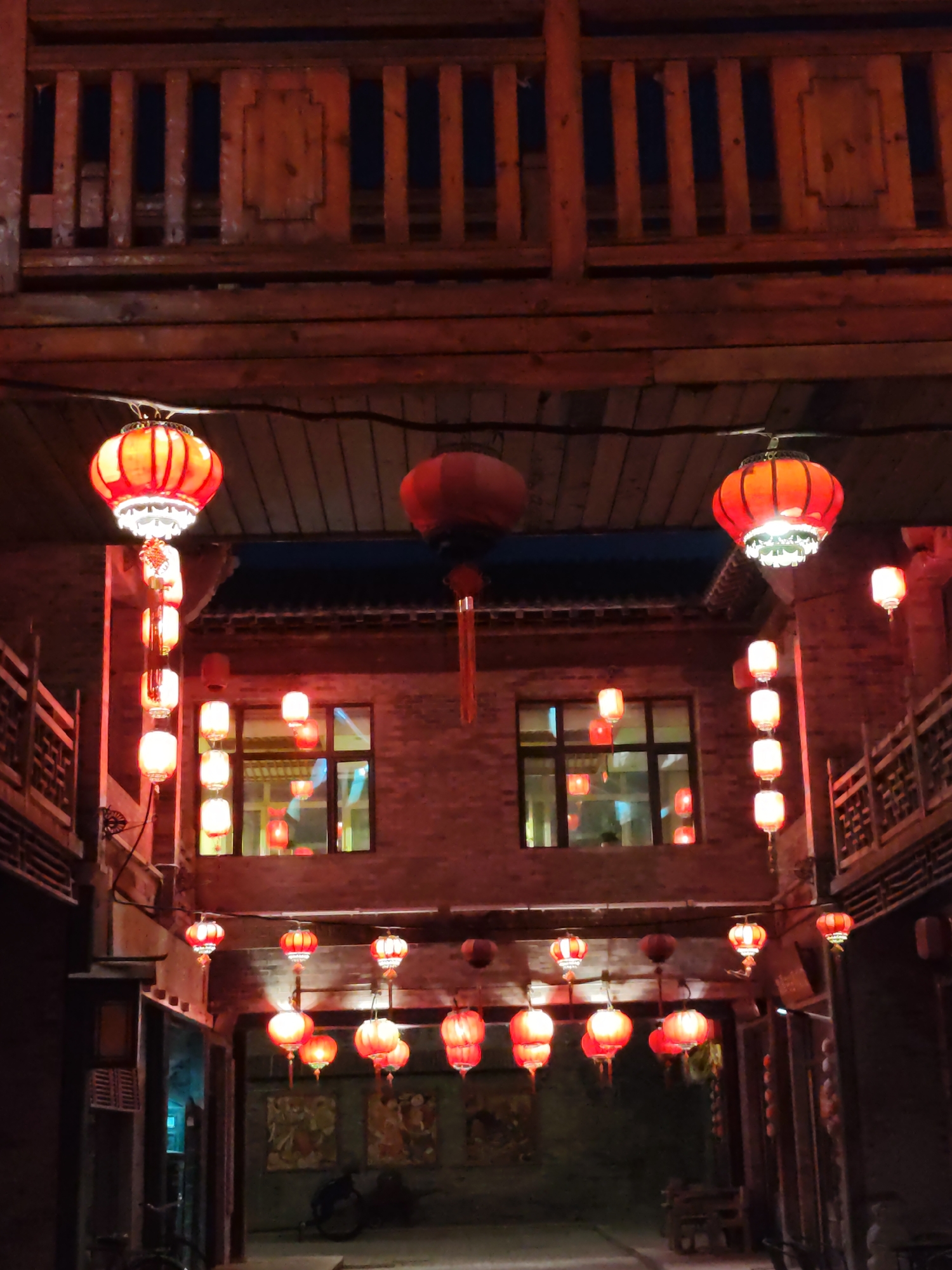 A nighttime scene depicts an alleyway in Lindian, China, adorned with numerous red lanterns. The lanterns, in various sizes and shapes, are suspended from wooden beams and lines strung across the alley. Some lanterns are illuminated, casting a warm glow on the brick buildings that flank the passageway. The buildings have traditional architectural elements, including wooden balconies and latticed windows. Faintly visible within the windows are reflections and hints of interior lighting. The ground of the alley appears to be paved. In the background, artwork or murals are affixed to the brick walls. Bicycles are parked along the sides of the alley. The overall atmosphere is festive and culturally rich, suggestive of a traditional festival or marketplace setting.