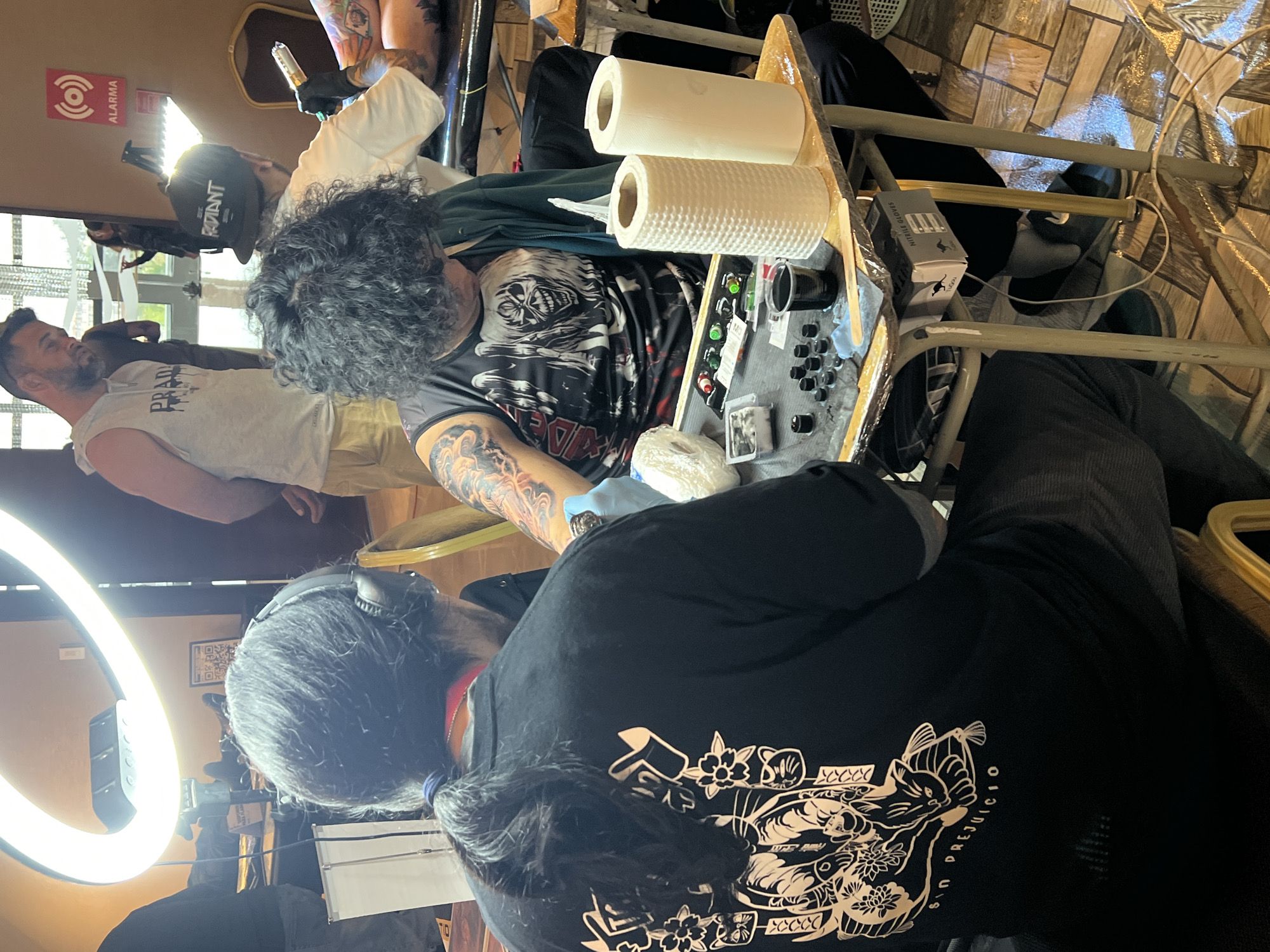 The image depicts an indoor scene, likely a tattoo convention or studio, with several individuals present. In the foreground, a person wearing headphones and a black t-shirt with a detailed white design on the back is seated, facing away from the camera. This individual appears to be engaged in a conversation or observation related to the activity occurring to their right.
To the right of the foreground subject, another individual is seated and actively tattooing the arm of a third person. This tattoo artist is wearing a black t-shirt featuring a graphic of a monster with sharp teeth and grey hair, and is wearing black nitrile gloves. They are using a tattoo gun and appear focused on their work. The client's arm, being tattooed, displays existing colorful tattoos.
A small table is positioned between these two individuals, holding various tattooing supplies including small ink caps filled with ink, a roll of plastic wrap, a box of nitrile gloves, and two rolls of paper towels. A bright ring light is visible in the upper portion of the image, illuminating the workspace.
In the background, several other people are visible, including a man wearing a grey sleeveless shirt and khaki shorts, observing the tattooing process. Another person, possibly a woman, is standing further in the background. The environment appears to be an indoor venue with tiled flooring and what looks like a draped table in the far background. A sign with a red circle and waves, labeled "ALARMA" (alarm in Spanish), is mounted on the wall. The overall atmosphere suggests a professional setting for tattoo art. While the image does not directly indicate the city of Arica, Chile, the presence of the Spanish word "ALARMA" and the context of a tattoo event common in various cities might imply a Latin American location.