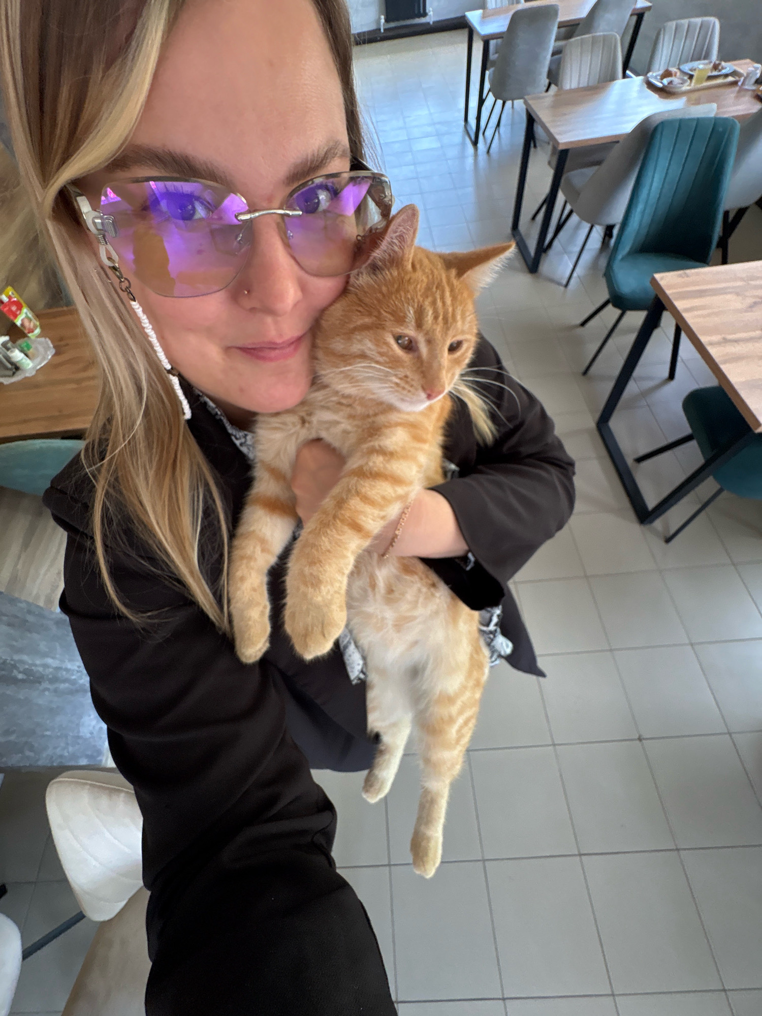 A woman with blonde hair and large, rimless glasses is holding an orange tabby cat. She is wearing a black jacket over a patterned shirt. The cat is being held against her chest. The background appears to be a restaurant or cafe with light-colored tiled floors and wooden tables and chairs. The image seems to be a selfie, as the woman is looking towards the camera. The location is not specified within the image.