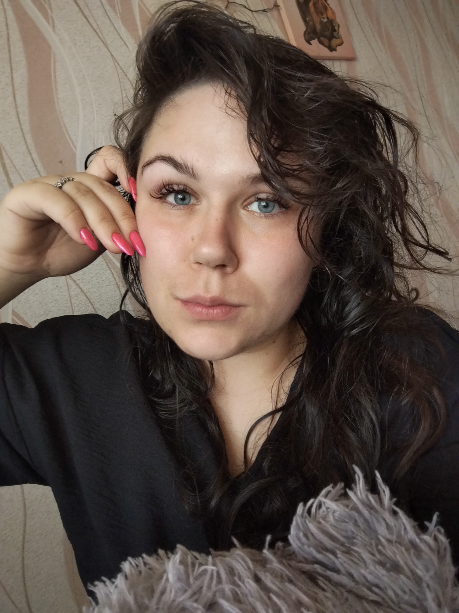 A woman is centered in the frame, gazing directly at the camera with a neutral expression. She has blue eyes, dark curly hair, and is wearing bright pink nail polish. Her left hand is raised near her face, with her fingertips touching her temple area. She's wearing a black top, and a fluffy grey material is visible at the bottom of the frame, possibly part of a blanket or garment. The background consists of a beige wall with a wavy pattern and a framed picture hanging above the woman's head. The image was captured indoors, seemingly in a domestic setting. There is no explicit evidence to confirm that the location is Chaikovsky, Russia.