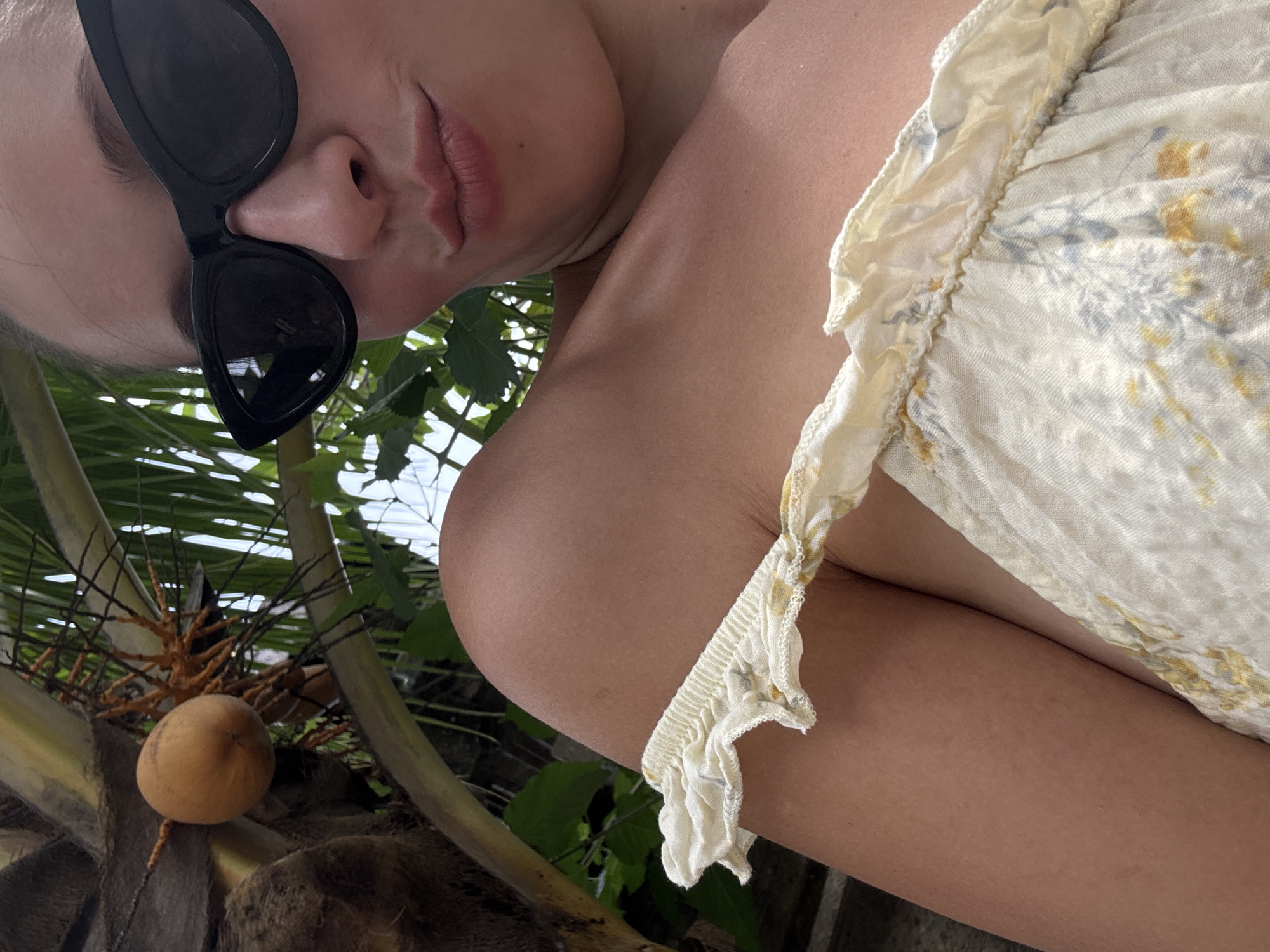 A close-up, angled shot shows a person with fair skin, wearing black sunglasses and a white, floral-patterned garment. The background includes palm trees with coconuts, suggesting a tropical setting. The location is likely North Kuta, Indonesia, based on the prevalent tropical vegetation and coastal setting.