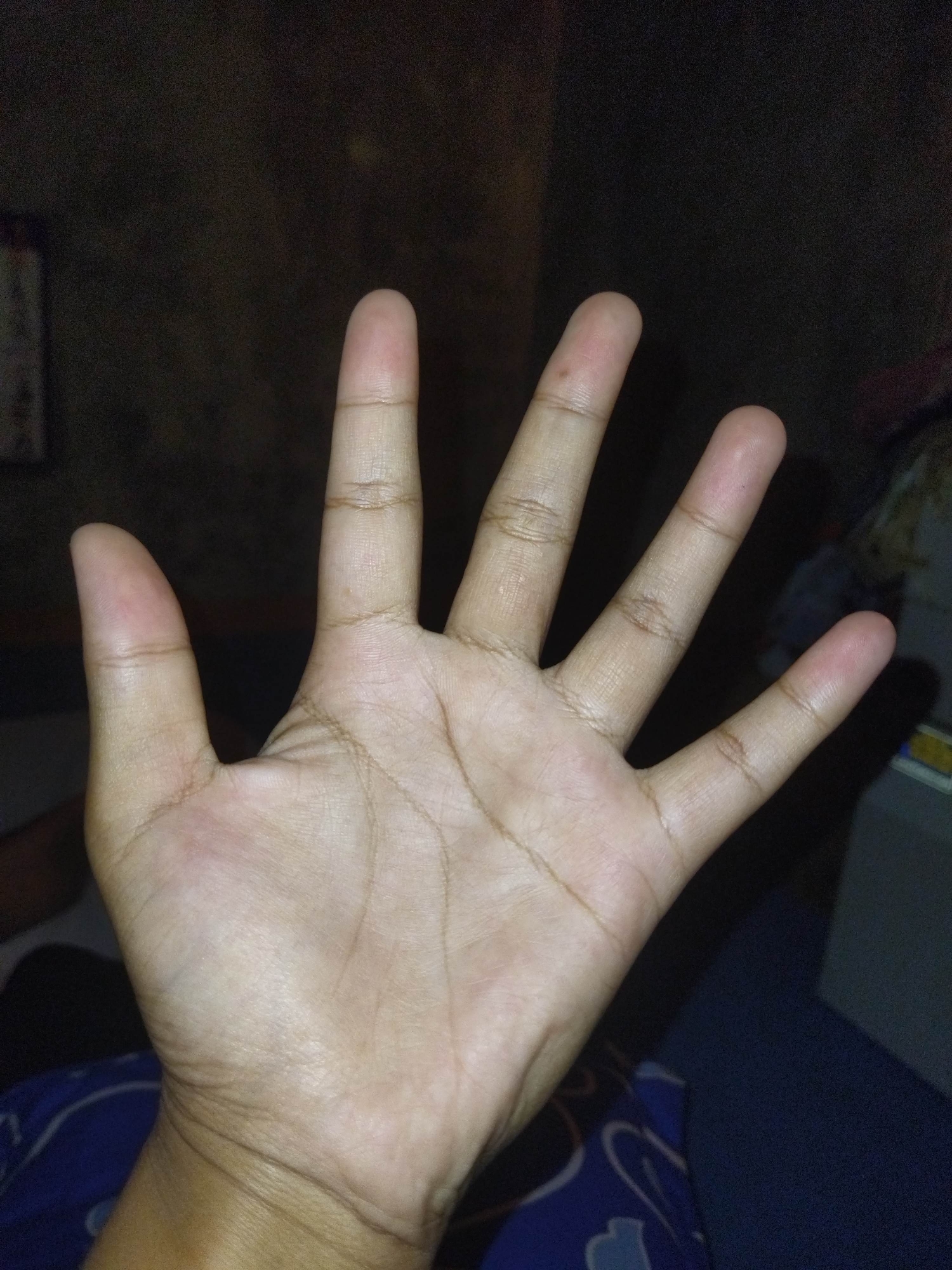 An adult's left hand is open, palm facing upwards, with fingers extended. The skin tone is light brown with visible lines on the palm and knuckles. The background is dark and out of focus, showing hints of dark fabric and possibly a wall. No identifiable landmarks for Suci, Indonesia are visible.