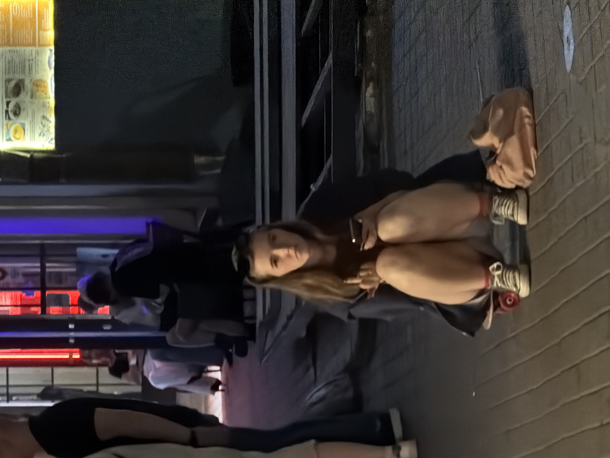 The image appears to be taken at night, likely in Moscow, Russia given the presence of signage in Cyrillic script. A woman is seated at an outdoor establishment. She's wearing sneakers and a dark garment. She is holding a device, possibly a phone or a digital device. A light-colored bag is near her feet. Several people are standing in the background. The area seems to be a public space possibly near restaurants or cafes.