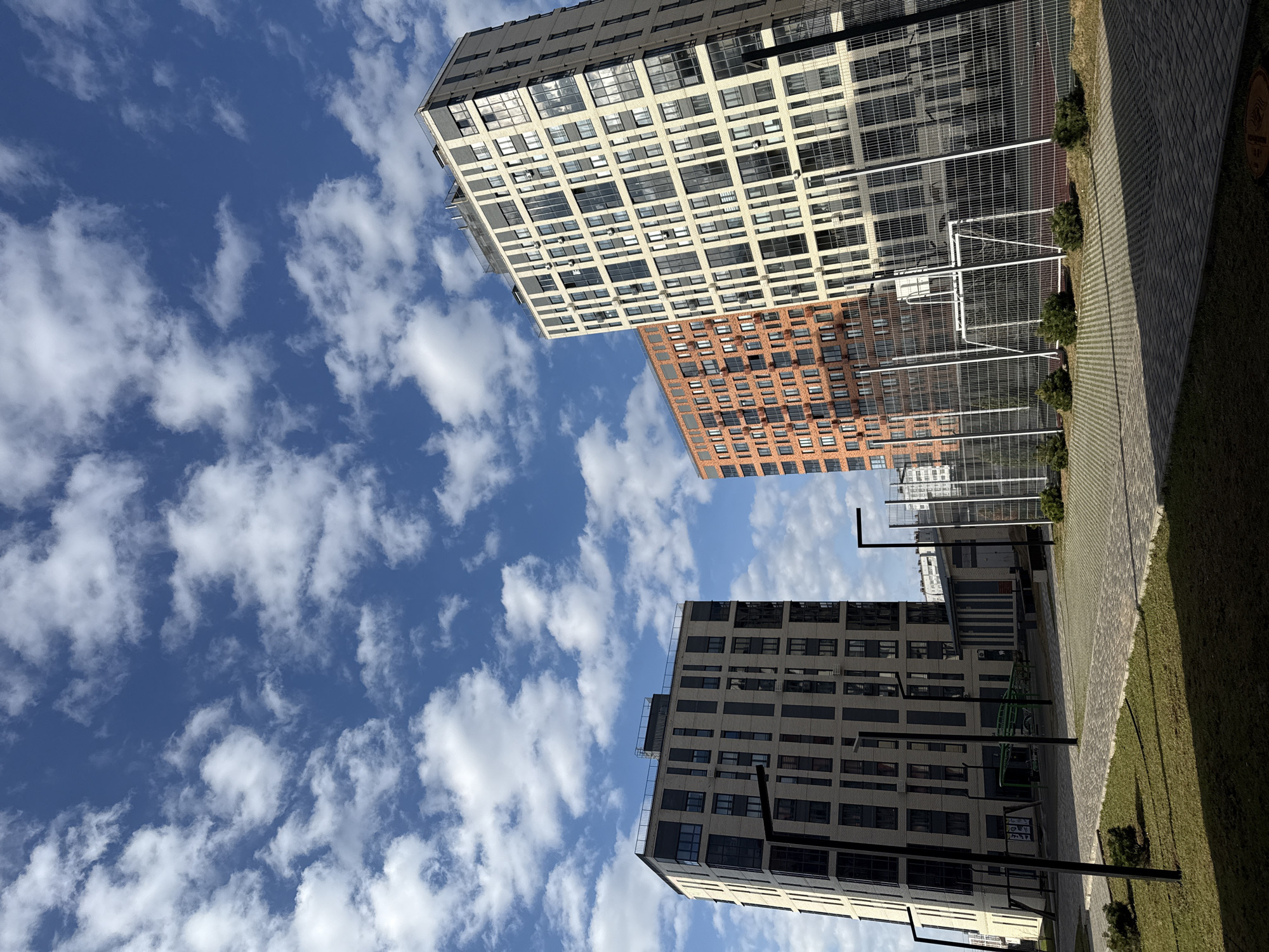 The image, taken in Izhevsk, Russia, presents a dynamic low-angle view of modern high-rise buildings against a bright blue sky filled with scattered white cumulus clouds. The scene is illuminated by clear daylight, suggesting it is either morning or afternoon.
Three multi-story buildings dominate the composition. The most prominent building, on the left, features a light-colored (beige or pale grey) facade with a grid pattern of numerous rectangular windows and some visible balconies. Behind it and slightly to the right, another tall building exhibits a distinctive reddish-brown brick facade, also with many windows. A third, similarly light-colored building is partially visible on the right side of the frame, appearing to be closer to the foreground.
The ground level, visible in the bottom right portion of the image, includes a paved walkway made of cobblestones or pavers, bordered by a strip of green grass. A white chain-link fence, possibly enclosing a sports court or recreational area, runs horizontally across the mid-ground. Small evergreen bushes are planted along this fence line. Several dark lampposts or streetlights are scattered in the foreground, and a green structure (possibly playground equipment or a bicycle rack) is visible behind the bottom right building.
There are no people present, and no specific activity is taking place, giving the scene a quiet, modern urban feel. On the paved walkway, a circular metallic utility cover or manhole is visible, but any text or symbols on it are too indistinct to be read.
