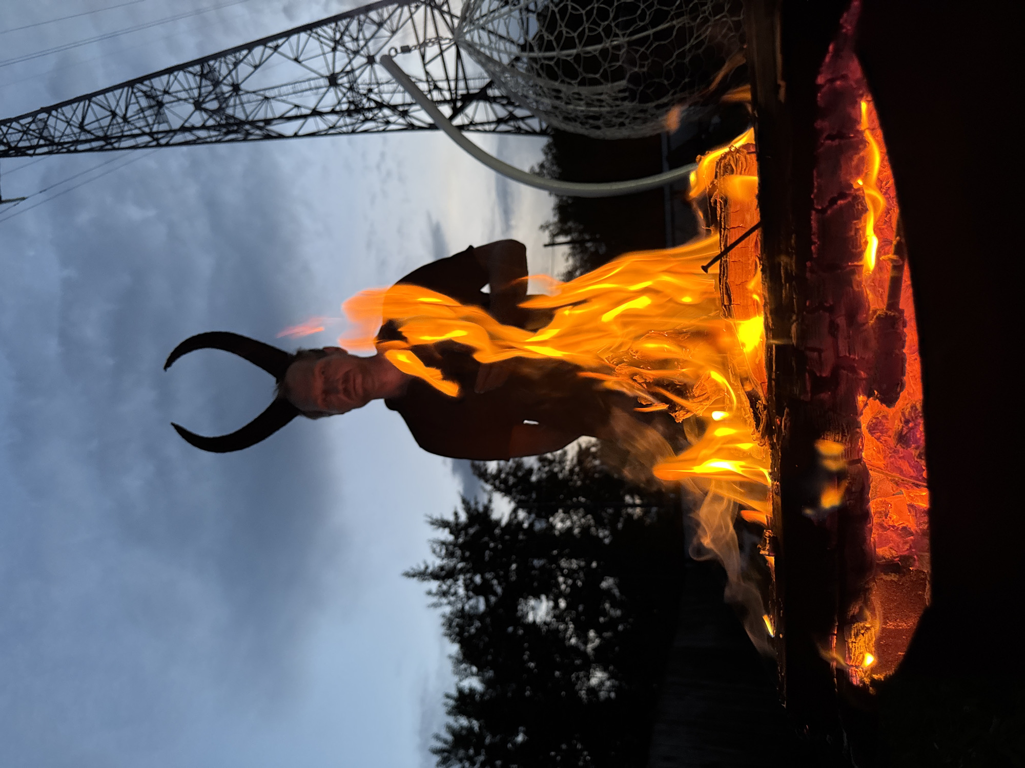 A man wearing large, dark, curved horns stands behind a bonfire, silhouetted against a twilight sky. The fire burns in a metal fire pit in the foreground, with bright orange flames and glowing embers. Behind the man, trees form a dark line against the sky, and a tall, lattice-like power pylon rises on the right side of the frame. A white, netted sphere hangs from a curved support to the right of the pylon. The scene appears to be outdoors, possibly in a backyard or a park in Chaikovsky, Russia, given the presence of the power pylon which is characteristic of industrial areas often found near cities. The overall mood is dark and somewhat dramatic, with the fire and the man's horns creating a striking contrast against the fading light.
