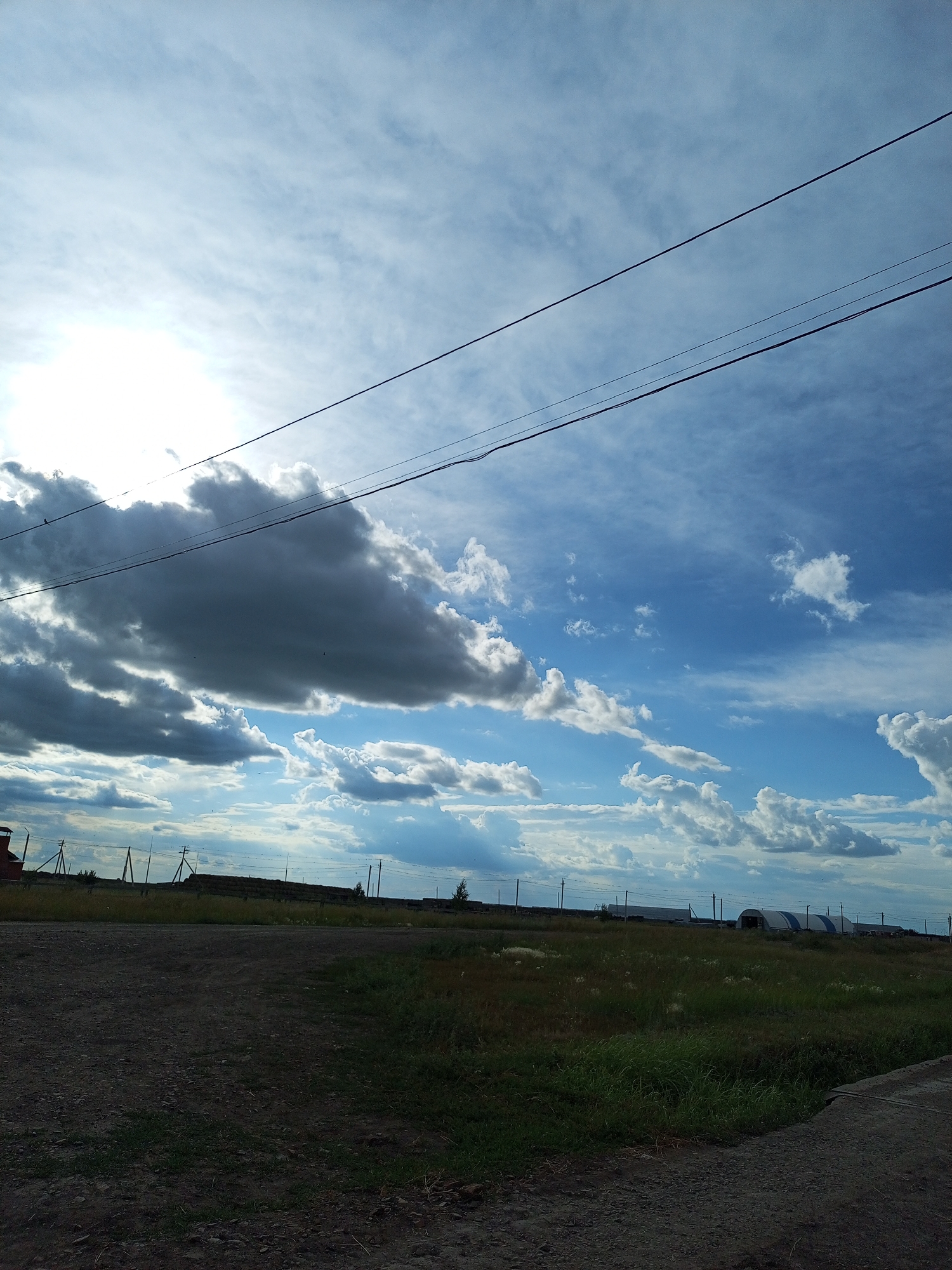 The image captures a view of the sky and a rural landscape, possibly on the outskirts of Burlí, Kazakhstan. The sky is a prominent feature, dominated by a bright sun shining through a layer of dramatic, dark grey clouds on the left, contrasted with patches of bright blue sky and scattered white cumulus clouds on the right. Several power lines traverse the upper portion of the image, running horizontally across the frame.
The foreground consists of a dirt or gravel road on the left and a grassy field on the right. In the middle ground, there is a distant line of hay bales, utility poles, and some low structures, including what appear to be agricultural buildings or greenhouses with curved roofs. A red brick structure, possibly a building or chimney, is visible on the far left. The overall impression is of an open, rural setting under a dynamic sky. There are no people visible in the image.