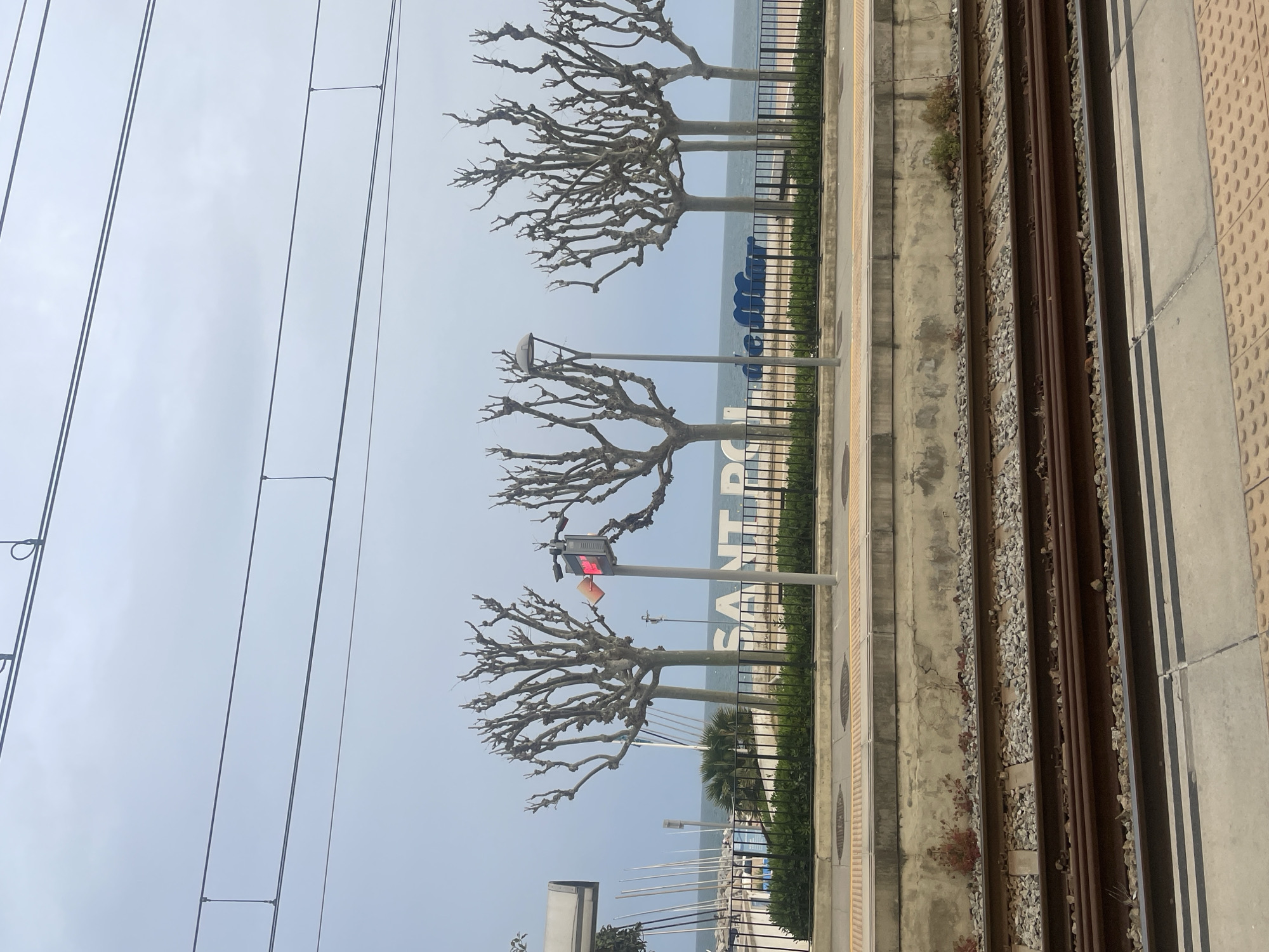 The image shows a view from a train platform in Sant Pol De Mar, Spain. The platform is on the right side of the frame, with textured, gray concrete slabs. To the left of the platform are train tracks, with visible gravel between the rails. Beyond the tracks, there's a concrete barrier followed by a decorative black metal fence. Trees with bare branches are planted along the fence line. In the background, the sea is visible with a large sign spelling out "SANT POL" in blue letters. A traffic light with a red light is positioned near the middle of the frame. Overhead electrical wires for the train run across the top left of the image against a pale blue sky. A few boats are visible in the water.