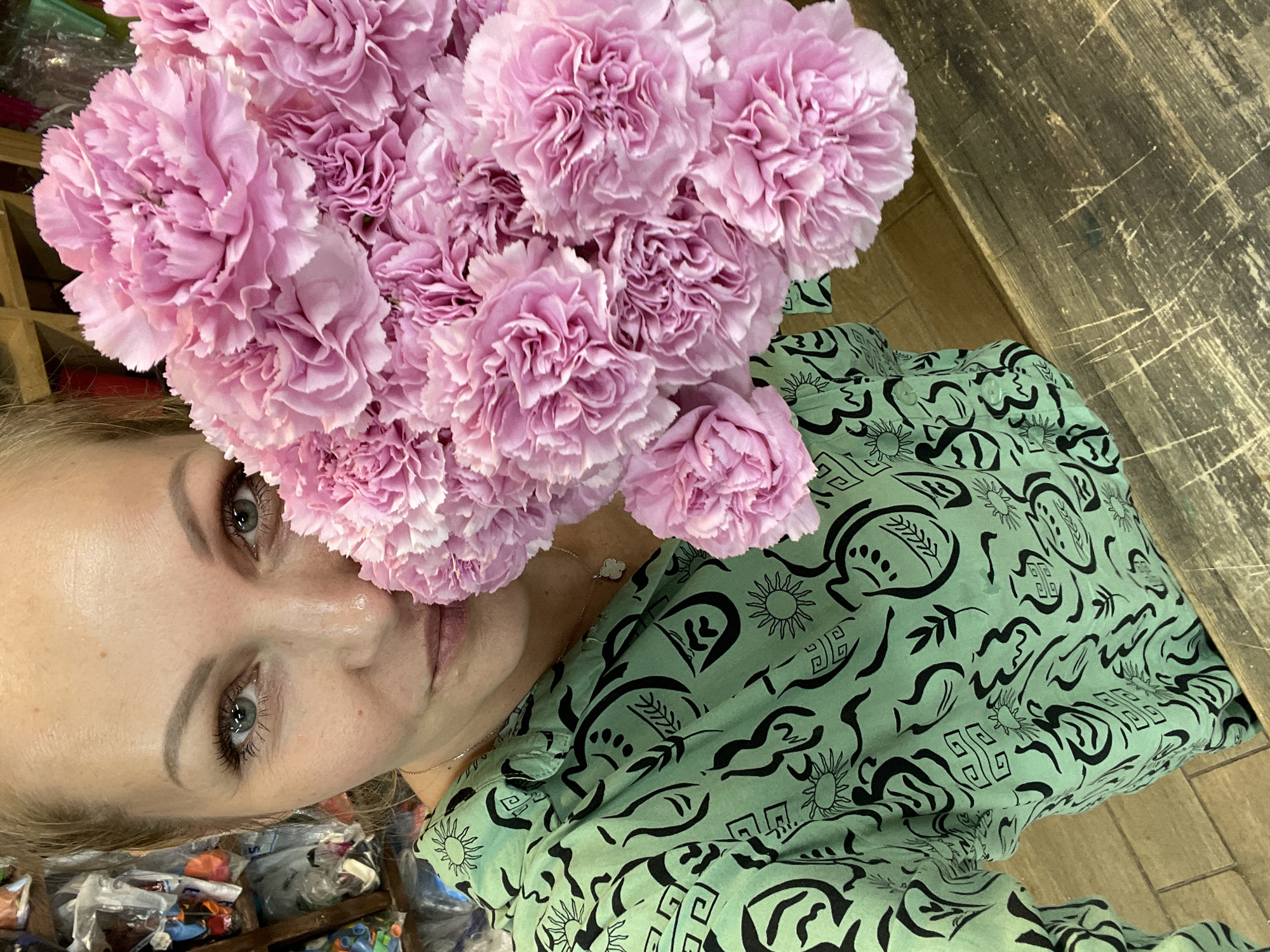 An adult Caucasian woman with blonde hair pulled back is holding a large bouquet of pink carnations. She is wearing a light green collared shirt with a black abstract pattern and a delicate necklace with a white pendant. The woman's face is partially obscured by the flowers, but her eyes, eyebrows, and part of her mouth are visible. The background appears to be an indoor setting, possibly a shop, with shelves of items visible on the left side and a wooden surface on the right. The woman is positioned in the foreground, looking towards the camera. The image was taken in the city of Kommunarka, Russia.