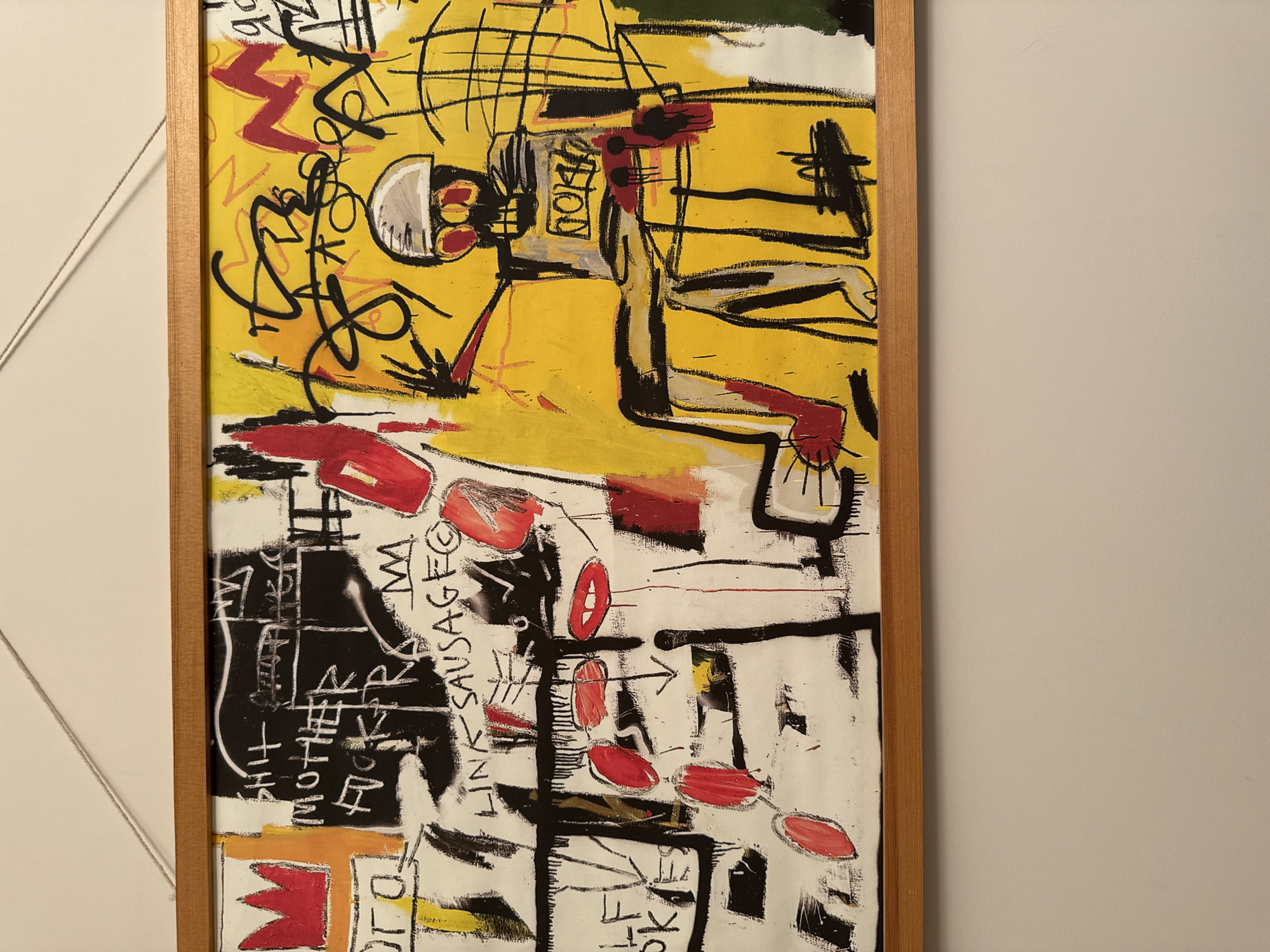 The image shows a framed painting with a mixed media style reminiscent of Jean-Michel Basquiat. The painting is divided into two distinct sections. The upper section features a dominant yellow background with abstract figures and scribbles in black, red, and white. There appears to be a stylized figure with linear body parts. The lower section has a white background with bold black lines creating a grid-like structure. Red oval shapes, seemingly random letters and words such as "SAUSAGE," "LINK," and "MOTHER," are incorporated into this section. The frame is a light-colored wood. The painting hangs on a light-colored wall. In the upper left corner, a small section of a white wall with a geometric pattern is visible. There is no indication that the painting is located in Moscow or Russia.