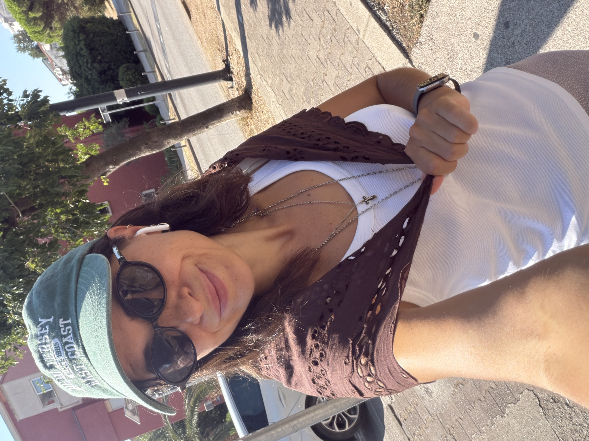 A woman with dark hair, wearing a green baseball cap, sunglasses, and a white tank top with a brown openwork scarf, stands on a paved sidewalk. She is holding a strand of her hair. A street with cars and trees lines the background. The scene is lit by bright sunlight. It's likely a street in Budva, Montenegro.