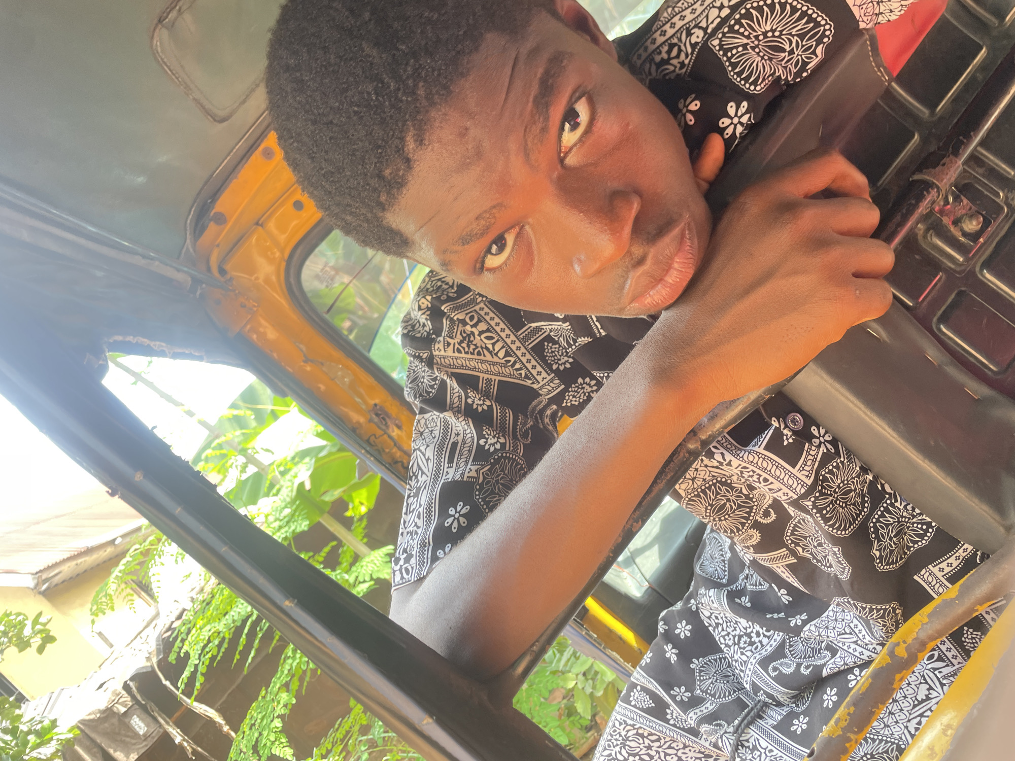 A young Black man with short, dark hair is the primary subject of the image, appearing inside a yellow and black vehicle. He wears a black and white patterned shirt. His head and hand rest on a portion of the vehicle's interior. The background shows green foliage and a building partially visible through the window. The photograph appears to be taken from a slightly rotated angle, looking upwards towards the subject. Based on visual cues and the location context of Nkpor, Nigeria, the vehicle may be a commercial tricycle, known as a "Keke" in Nigeria. The photograph was taken during daylight, as evidenced by the natural lighting.
