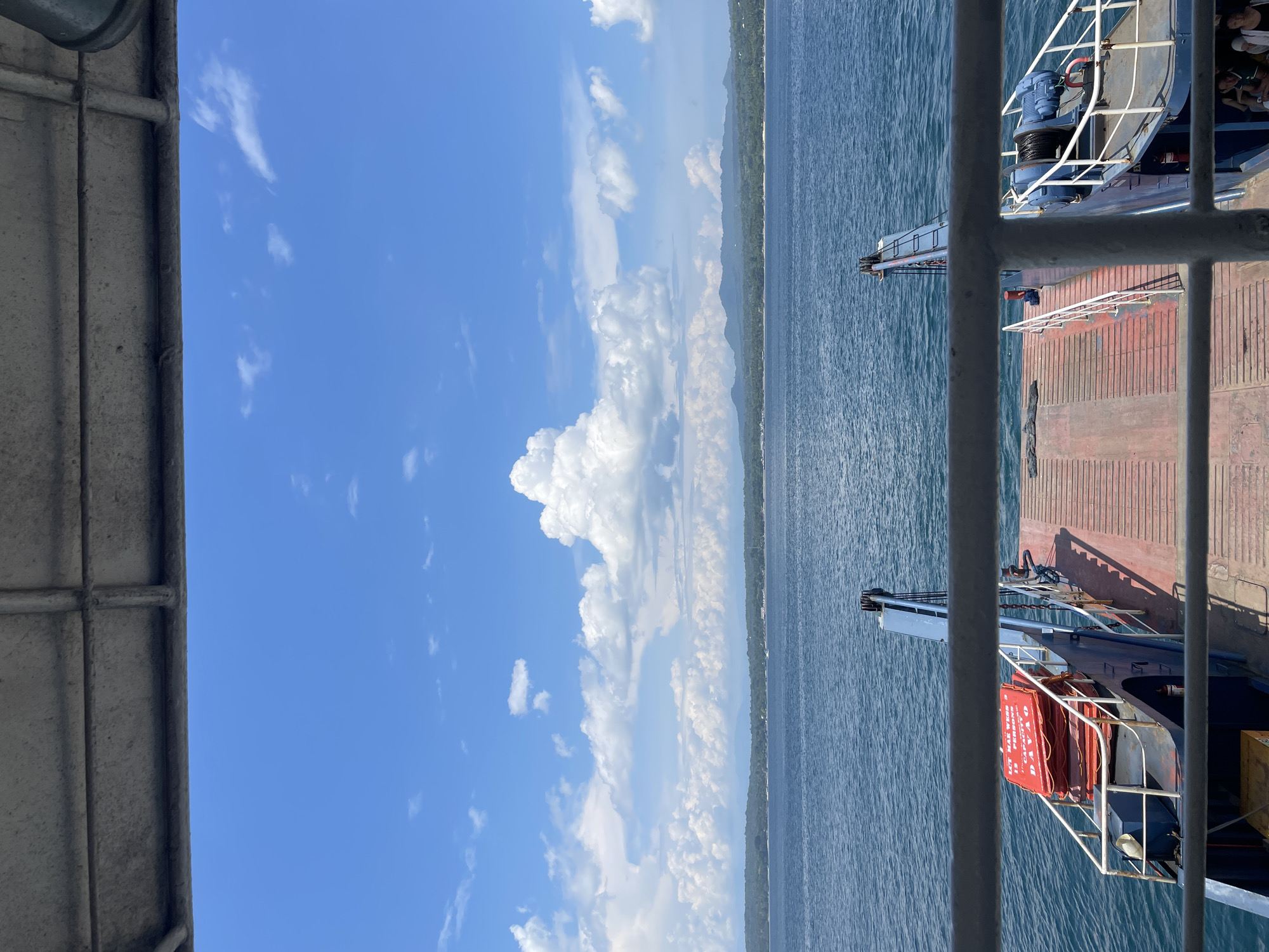 The image captures a view from the deck of a ferry, looking out over the Davao Gulf towards the coast of Davao City, Philippines. The sky is a vibrant blue, adorned with large, fluffy white clouds. The sea is a deep blue, with gentle waves. In the foreground, the ferry's wooden deck and metal railings are visible, along with life vests stacked on a platform. The coastline in the distance is lined with lush green trees and mountains, suggesting a tropical landscape. The overall scene conveys a sense of travel and scenic beauty, characteristic of a ferry ride in the Philippines.