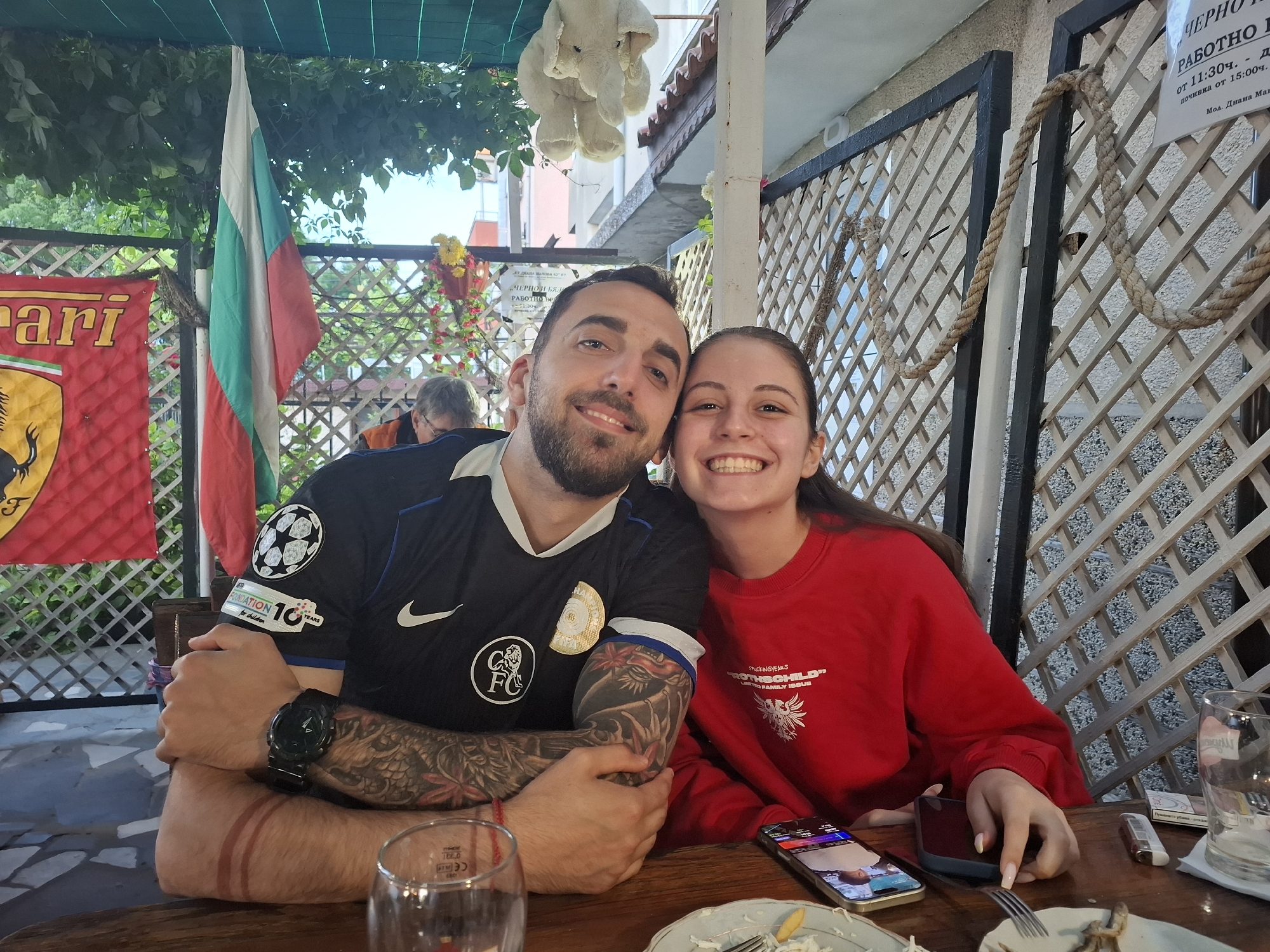 The image depicts a man and a woman seated at a wooden table, likely at an outdoor restaurant or cafe in Tsarevo, Bulgaria. They are smiling and posing closely together, suggesting a friendly or romantic relationship. The man is wearing a black football jersey with various patches and tattoos on his arm, while the woman is wearing a red sweatshirt. In front of them are plates with remnants of a meal, a smartphone, and glasses. In the background, there's a Bulgarian flag, a Ferrari banner, decorative plants, and a lattice wall. A sign with Bulgarian text is visible on the right side of the image, which likely indicates the working hours. An elephant plush is hanging from the ceiling. The scene conveys a relaxed and social atmosphere.