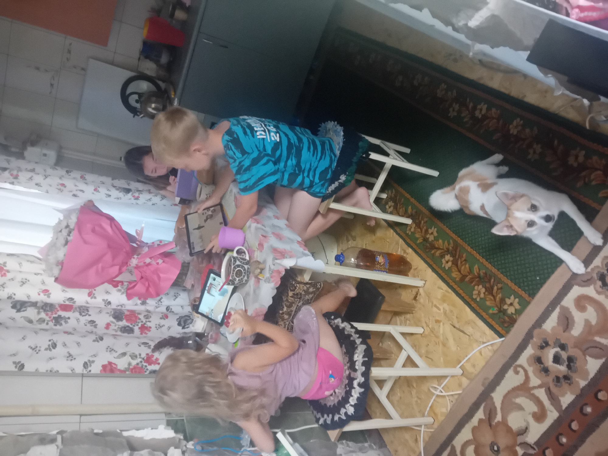 In a kitchen in Chuhuiv, Ukraine, three children and a dog are gathered around a table. A young girl with blonde hair sits on a stool, facing away from the camera, and is holding a tablet. A boy with blonde hair, wearing a blue t-shirt with a black pattern, is seated at the table, also looking at a tablet. To the right, a girl with dark hair is partially visible, looking down at a tablet. The table is covered with a floral tablecloth and has several items on it, including a mug, a bowl, and a bottle of soda. A bouquet of pink flowers is positioned behind the table. In the foreground, a small dog with white and brown markings lies on a rug. The kitchen appears to be in a state of disrepair, with exposed walls and a rustic decor. The overall scene depicts a casual moment of children engaged with technology in a home environment.