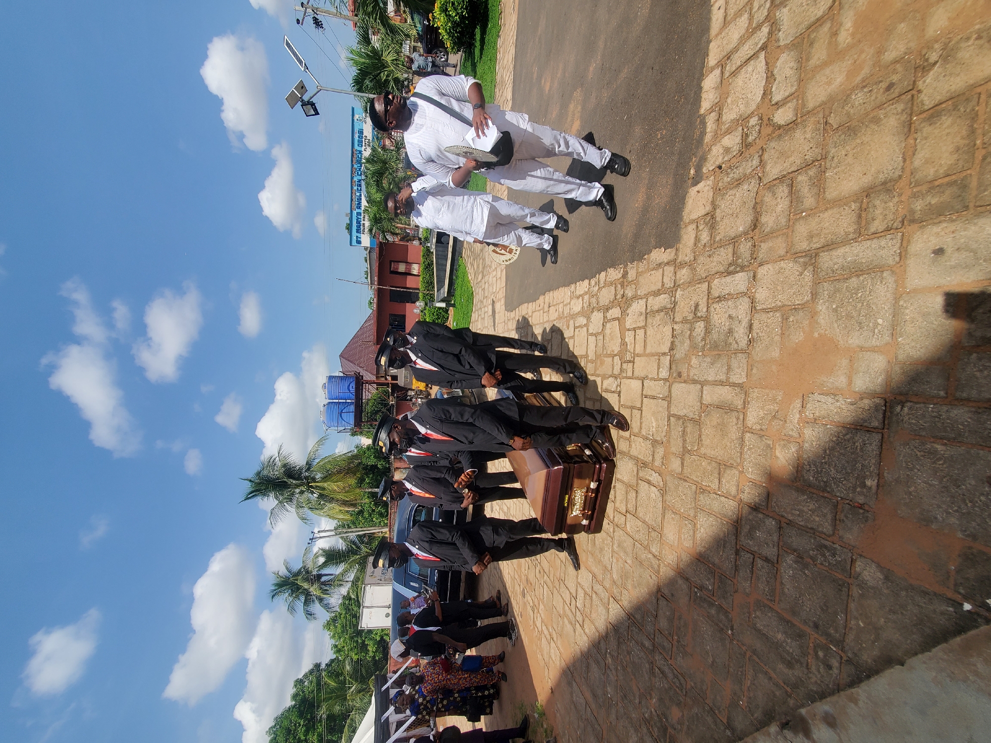 The image captures a funeral procession in Nkpor, Nigeria. Pallbearers dressed in black suits and hats carry a casket. Two men in white outfits are walking next to them. A group of mourners is visible in the background. The procession is taking place on a brick-paved area near a building. Above are sunny skies with clouds. In the background is a church building.