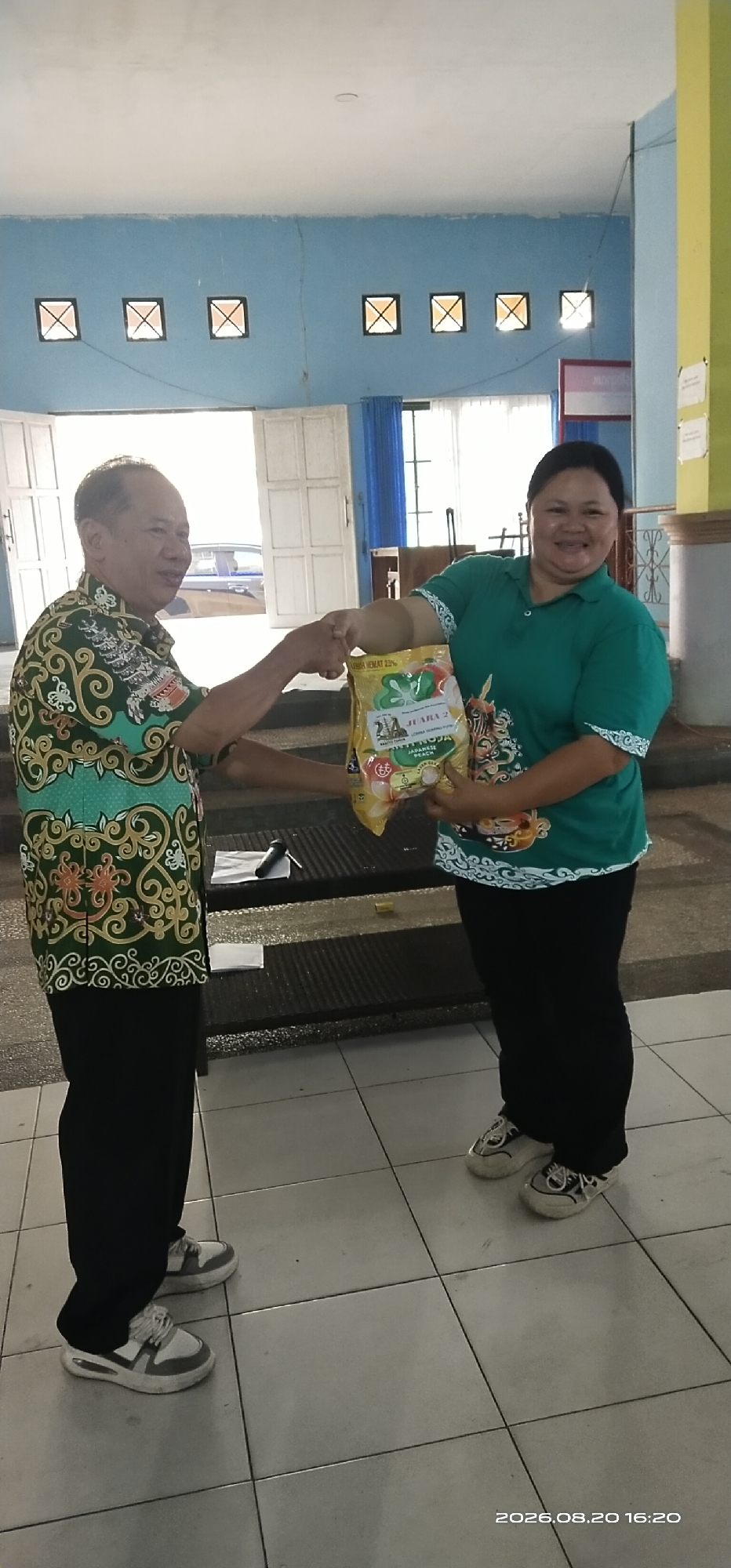 In Tamiang Layang, Indonesia, two individuals are captured in an indoor setting. On the left, a smiling man in a patterned green shirt and black pants extends his hand, appearing to be handing a yellow bag of snacks to a woman. The woman, on the right, is wearing a green polo shirt with white trim and black pants. She is also smiling and holding the bag. The bag features text and images of fruit. The background shows a blue wall with square vents, white doors, and a section of yellow wall. The floor is tiled in a light color. A timestamp at the bottom right of the image reads "2026.08.20 16:20".